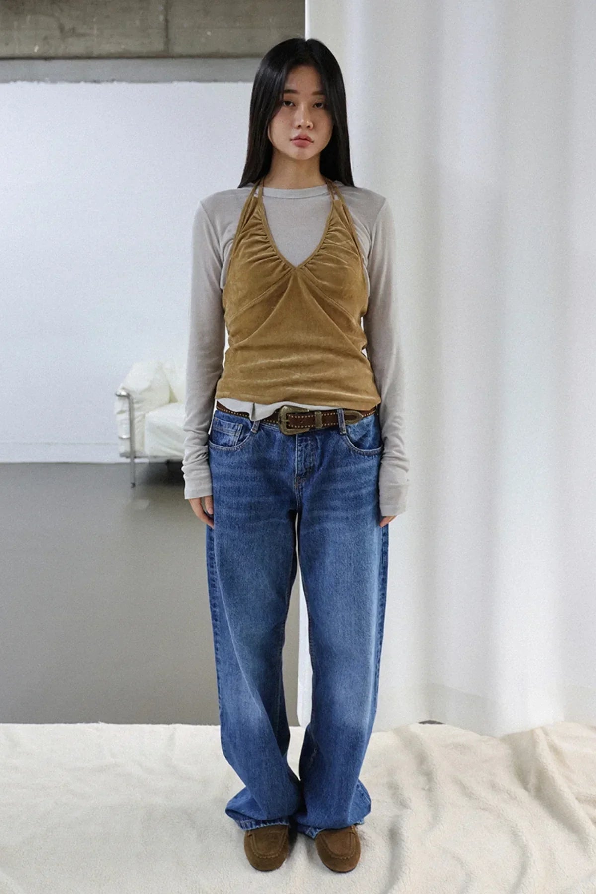 Young woman wearing camel suede shirring bra top over beige long sleeve shirt with blue jeans and brown shoes in minimalist room