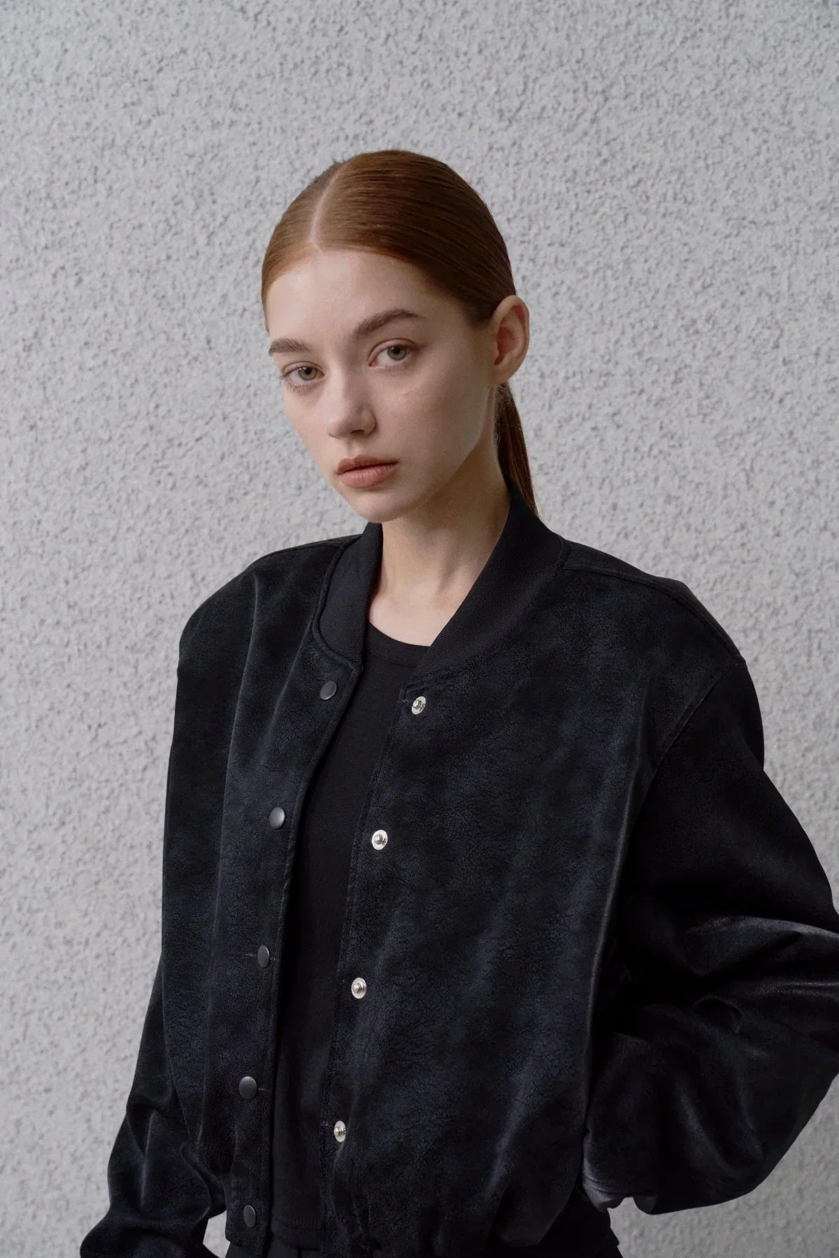Woman wearing a black suede stadium jacket with snap buttons against a textured gray wall