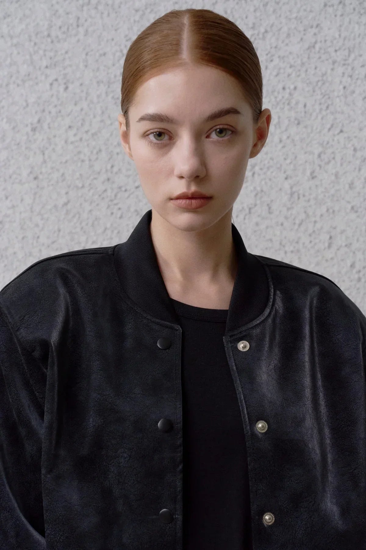 Close-up of young woman with slicked-back hair wearing black suede stadium jacket with snap buttons