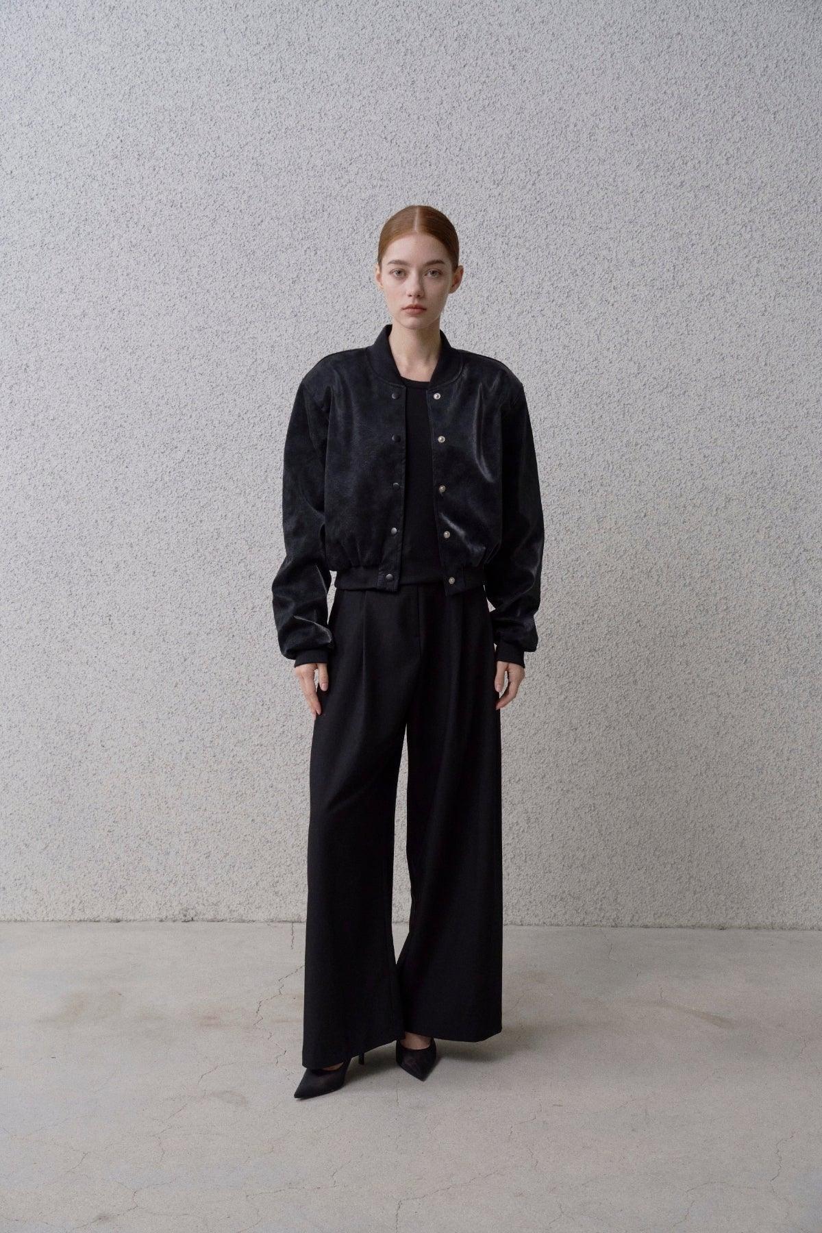 Woman wearing black suede stadium jacket, wide-leg pants, and heels against gray textured wall