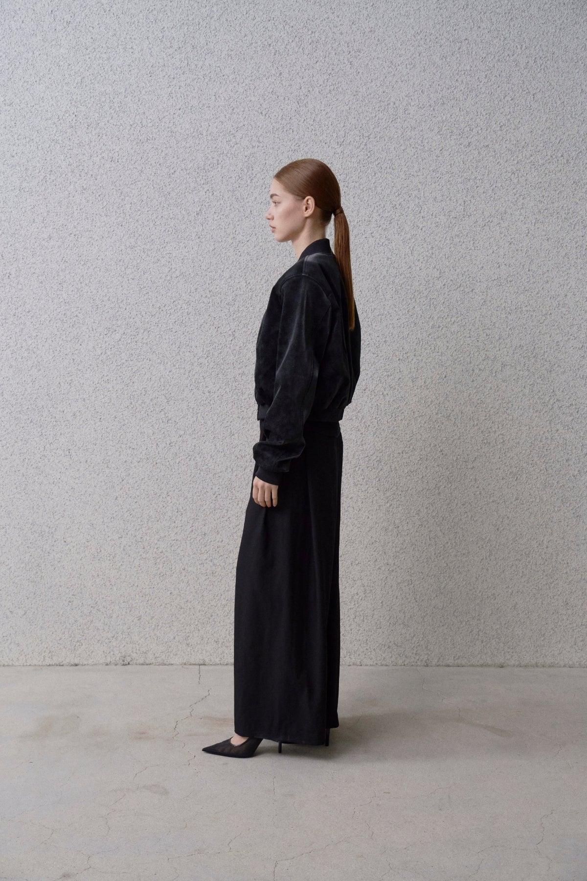 Side view of woman with ponytail wearing a black suede stadium jacket, long black skirt, and black heels against a textured wall