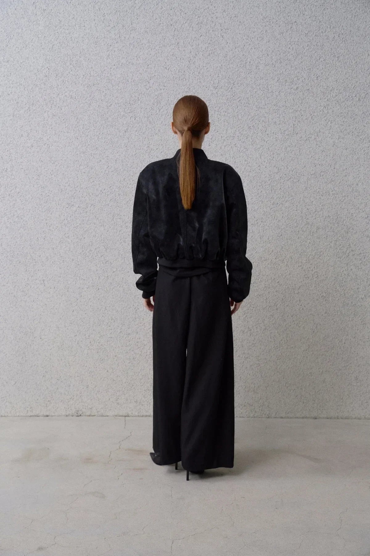 Back view of a woman wearing a black suede stadium jacket and wide-leg black pants in minimalist setting