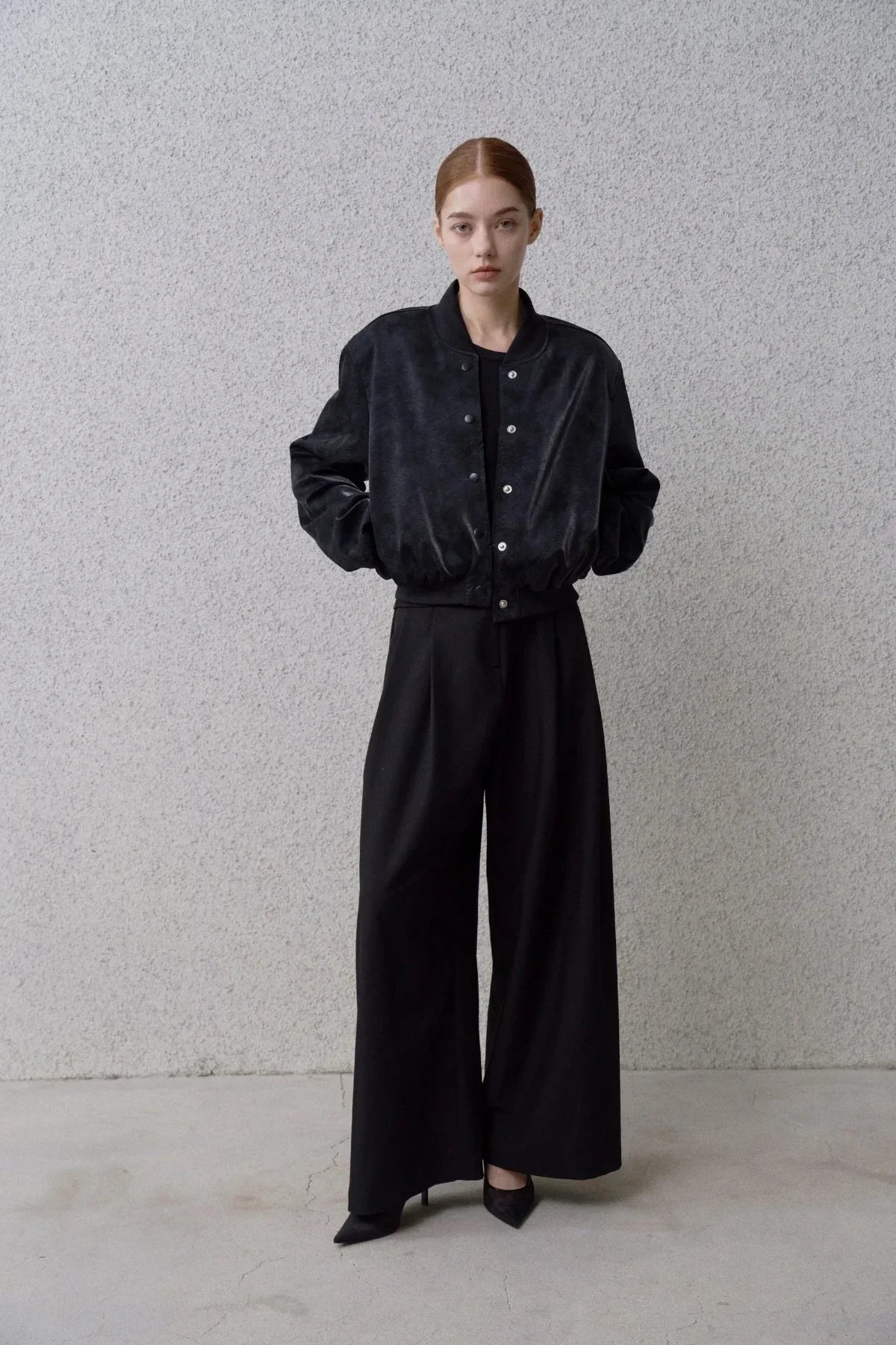 Model wearing black suede stadium jacket with snap buttons and wide-leg black pants in studio