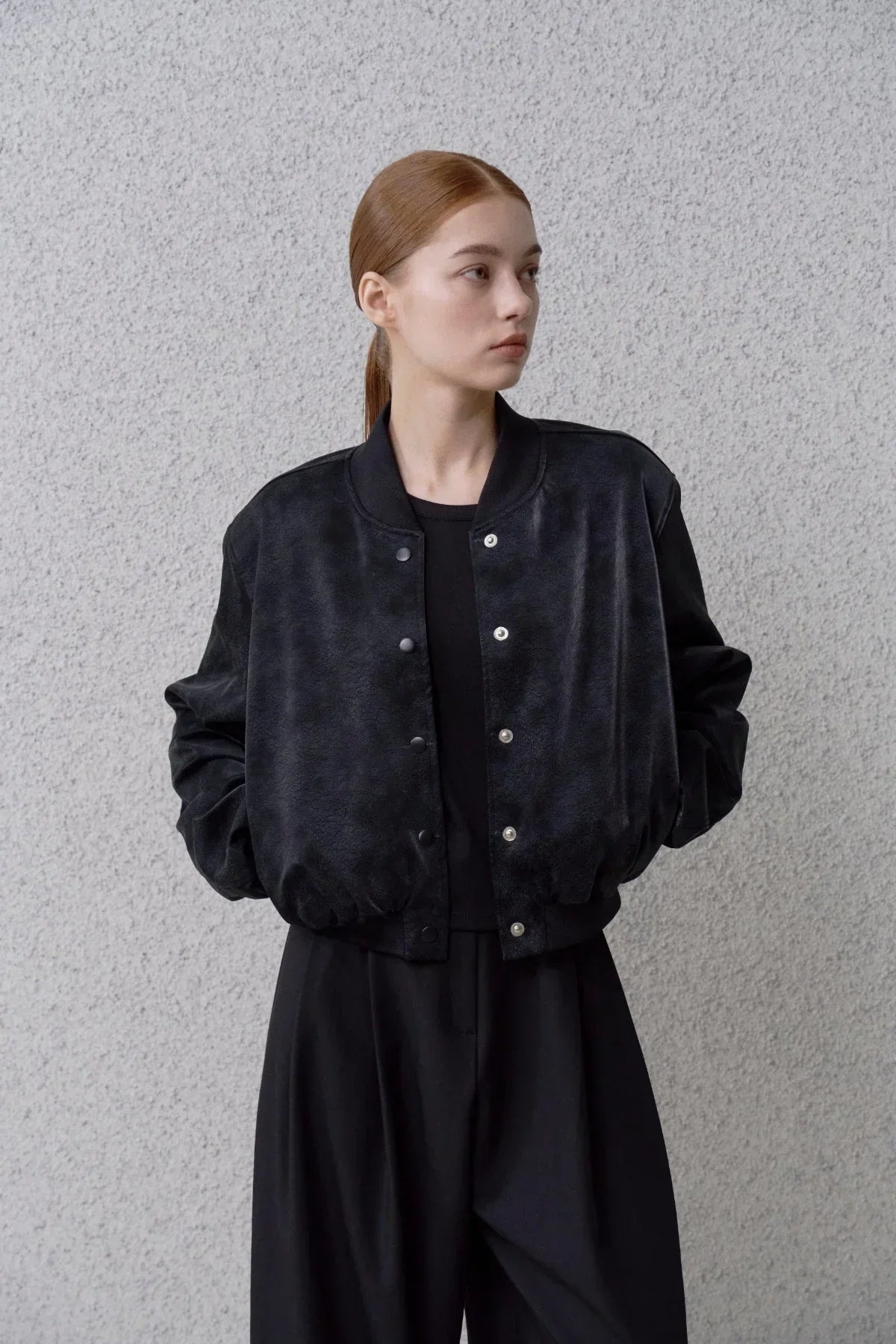 Young woman wearing black suede stadium jacket with snap buttons, black pants, standing against gray textured wall