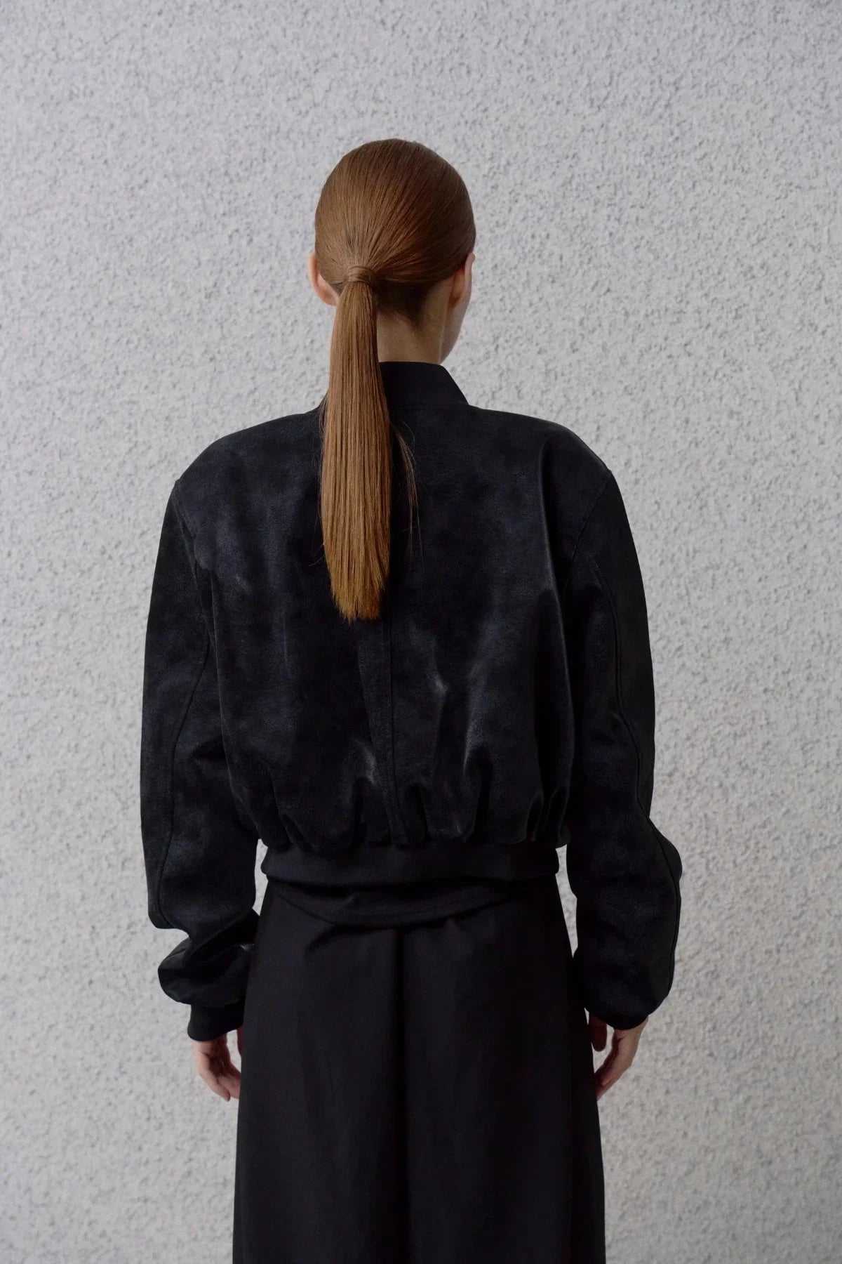 Back view of a woman with a sleek ponytail wearing a black suede bomber jacket against a textured gray wall