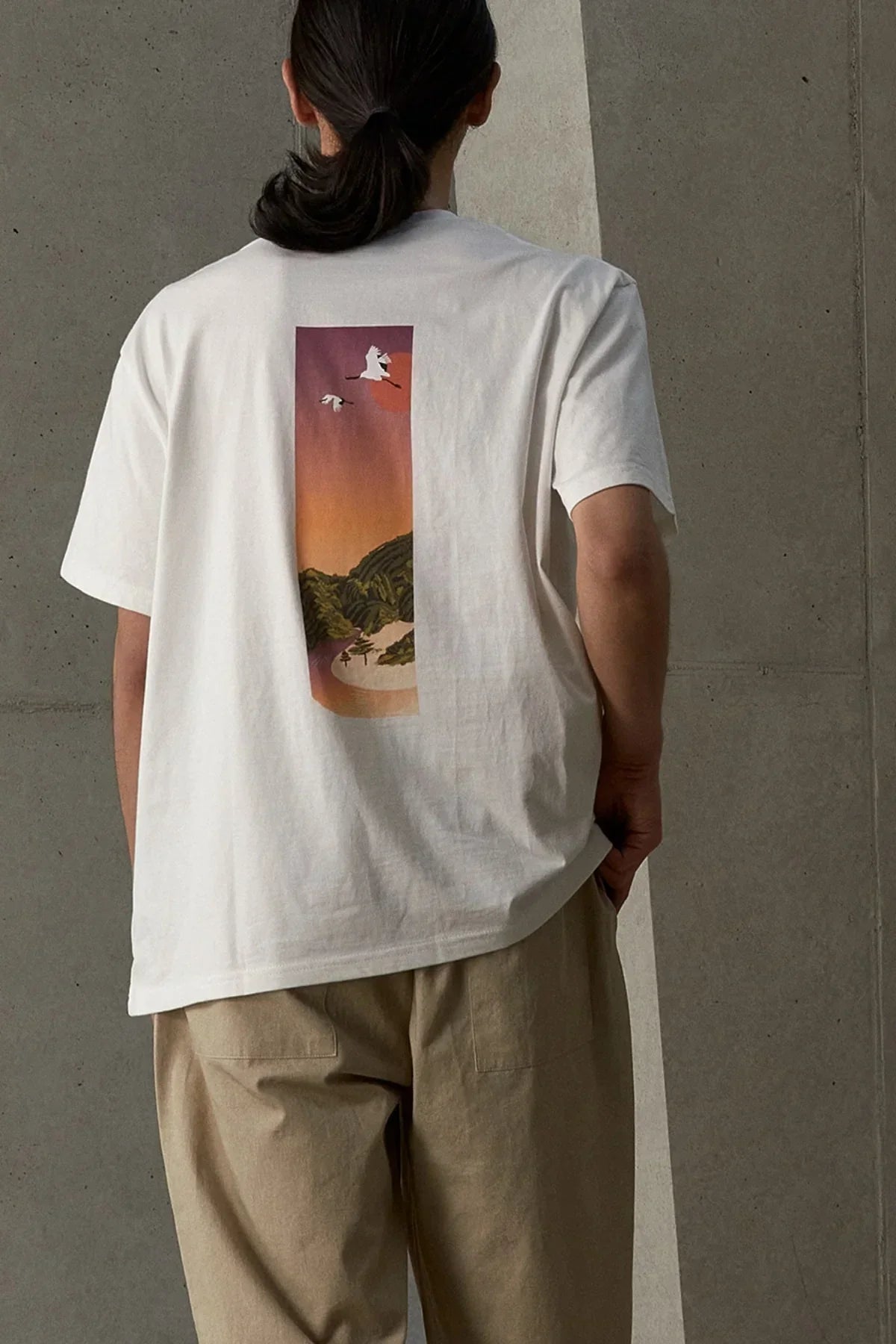 White t-shirt with colorful sunset and crane birds graphic on back, worn by person in beige pants