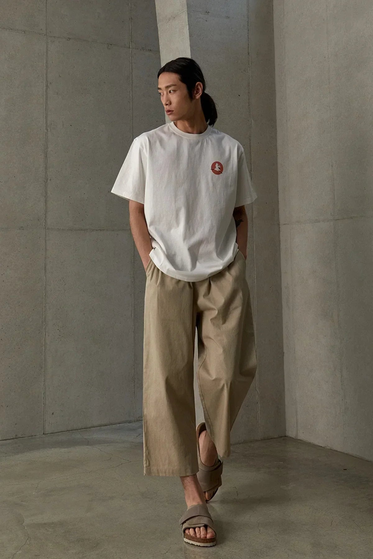 Man wearing white graphic t-shirt, beige wide-leg pants, and sandals in minimal concrete room