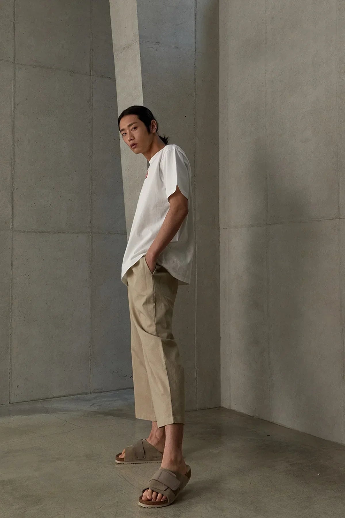 Young male model in white t-shirt, beige cropped pants, and brown sandals standing against concrete wall