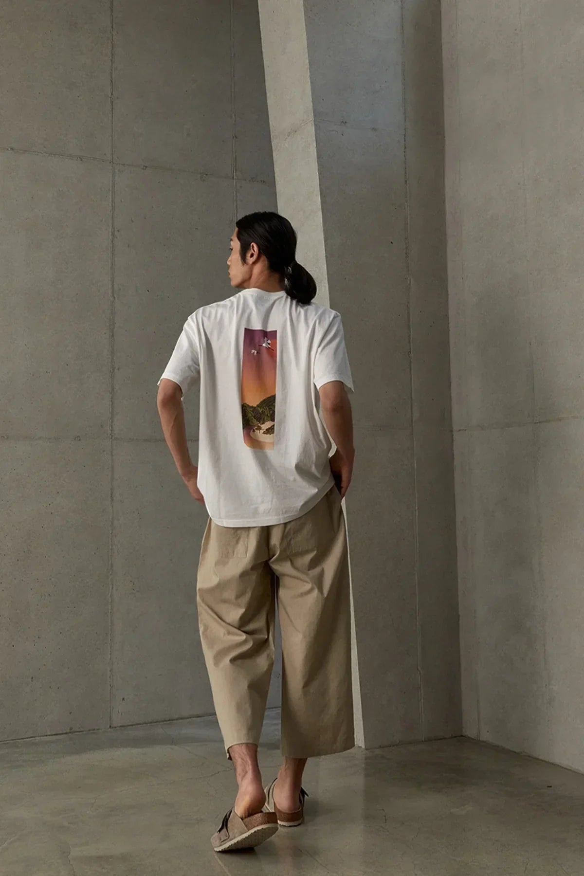 Man wearing white t-shirt with sunset beach graphic on back, beige wide pants, in minimalist concrete room