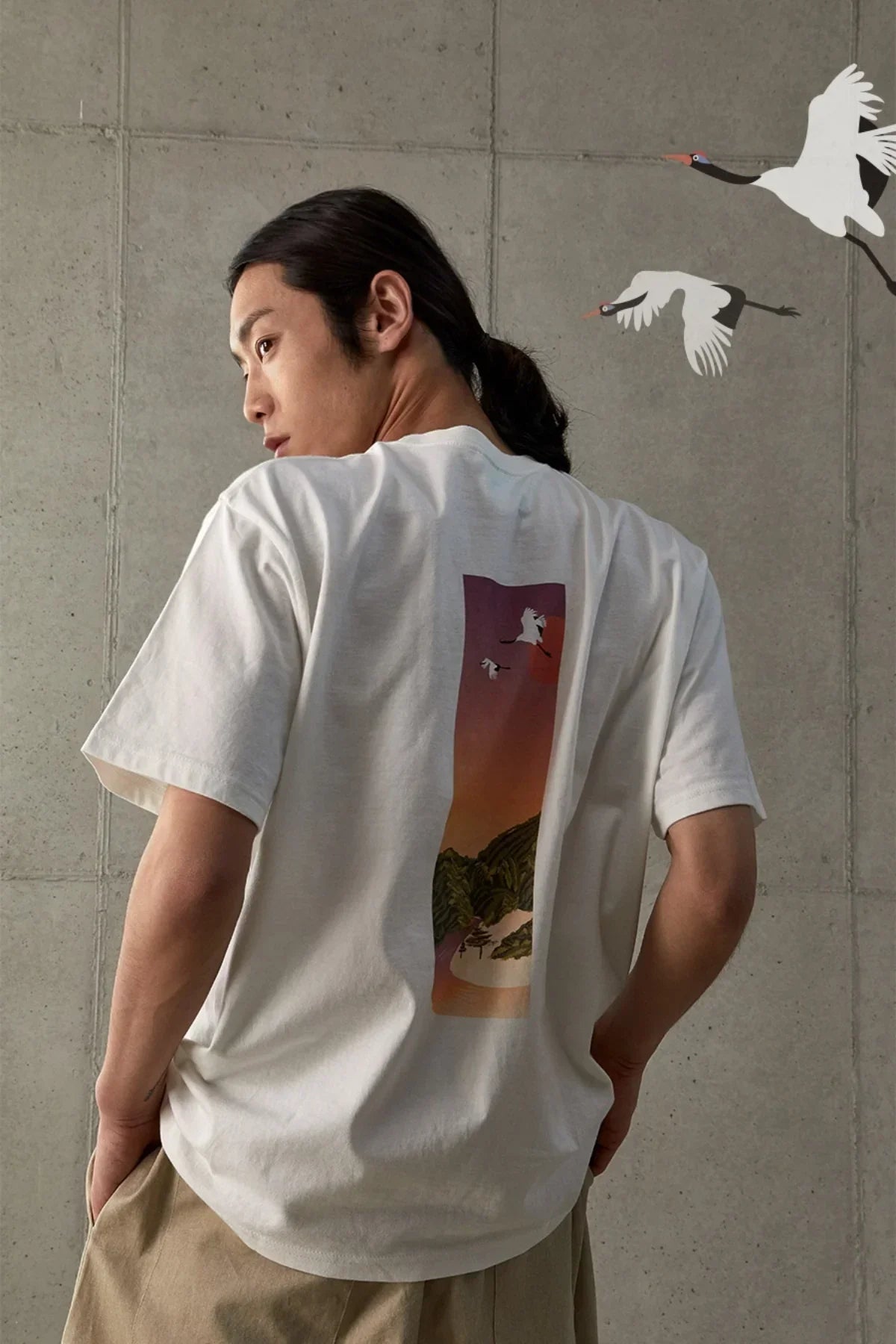 Man wearing white t-shirt with sunset and flying cranes design, standing against concrete wall
