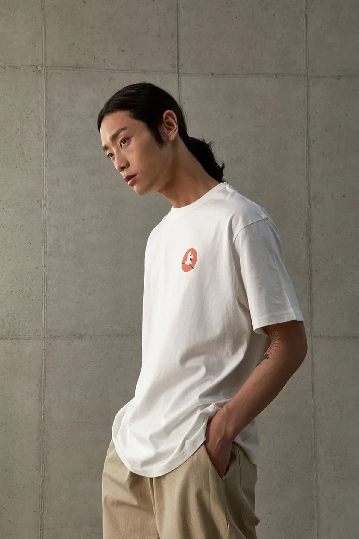 Young man wearing white graphic t-shirt with orange circle design and beige pants against concrete wall