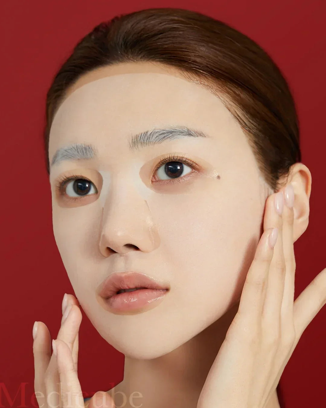 Close-up of woman applying white sheet mask on face with red background, skincare beauty routine