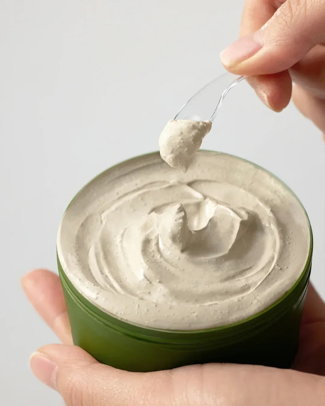 Hand holding green jar of creamy beige clay mask with small spoon scooping texture
