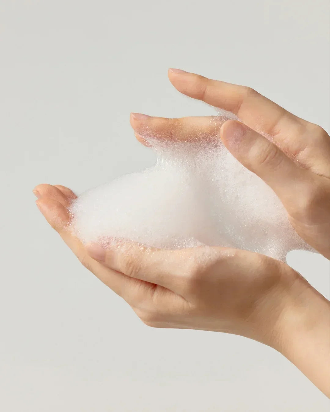 Hands holding thick white foam cleanser against a plain light background