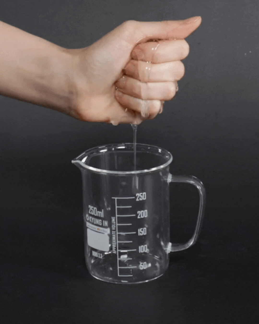 Hand squeezing clear serum into 250ml glass measuring cup against black background