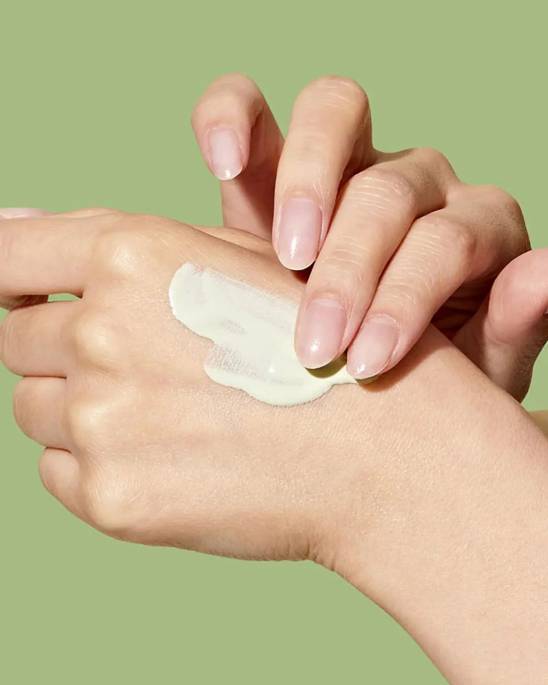 Close-up of hands applying creamy white skincare product on hand against green background