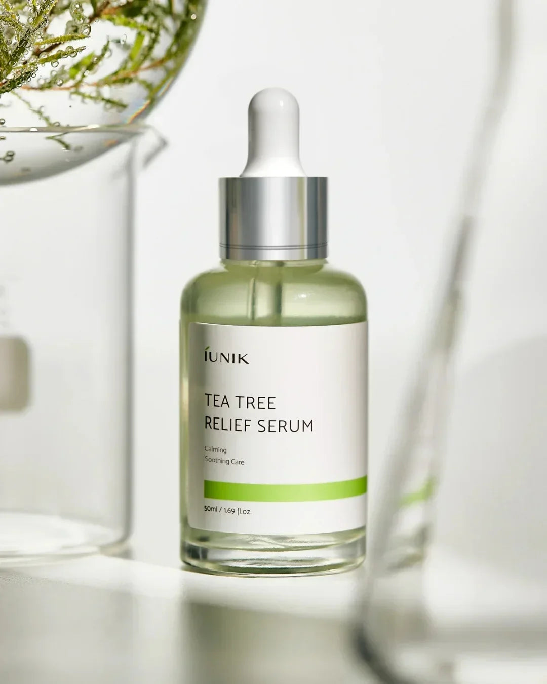 iUNIK Tea Tree Relief Serum bottle with dropper on white surface with glass and green plant