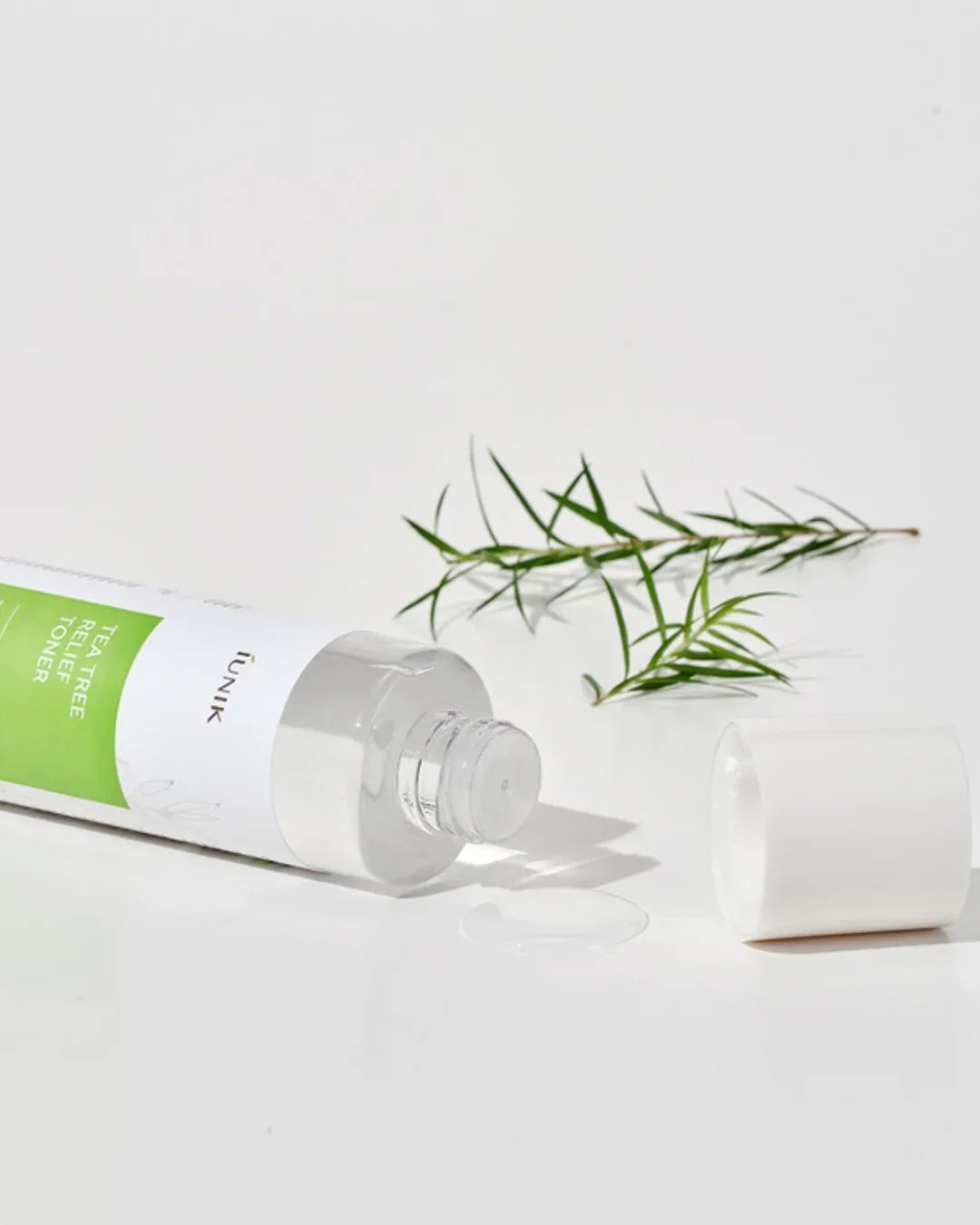 iUNIK Tea Tree Relief Toner bottle lying on white surface with green tea leaves and clear liquid