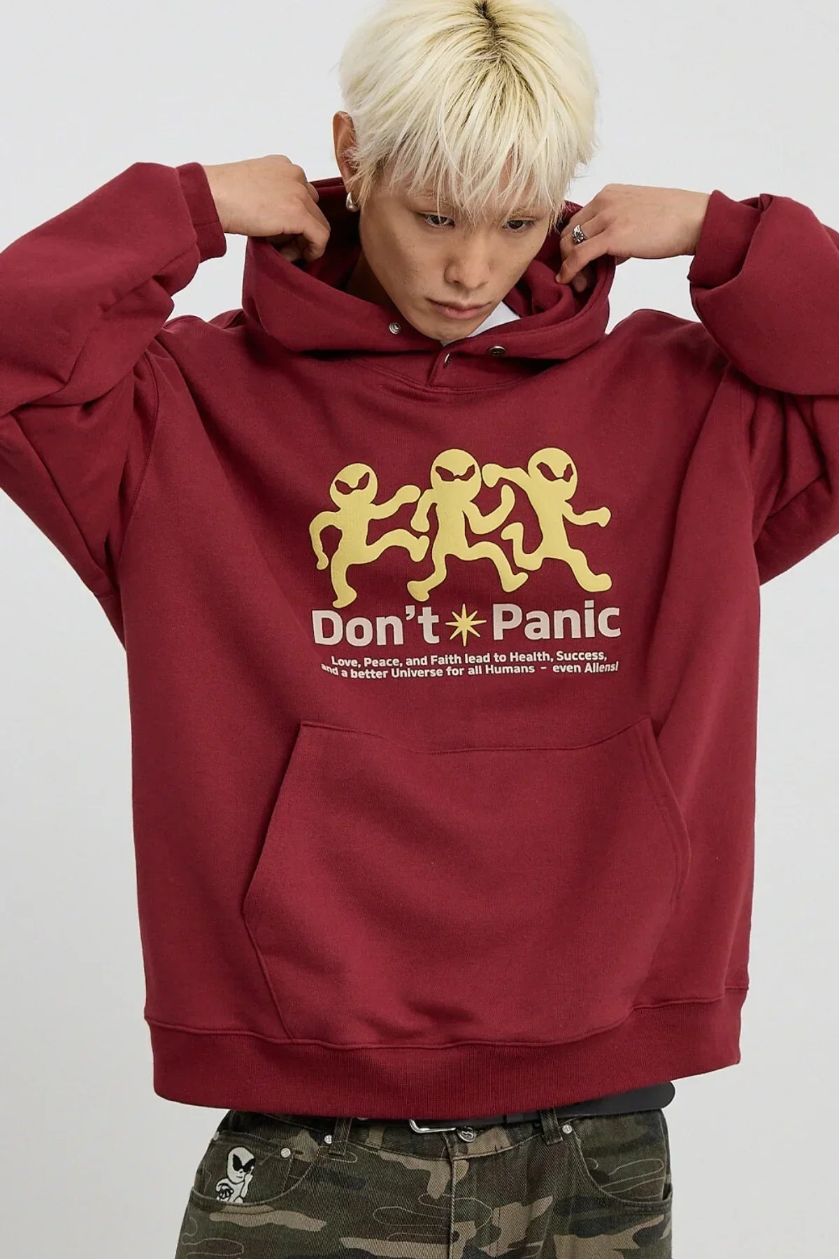 Blond man wearing burgundy hoodie with dancing alien graphic and text 'Don't Panic', camo pants