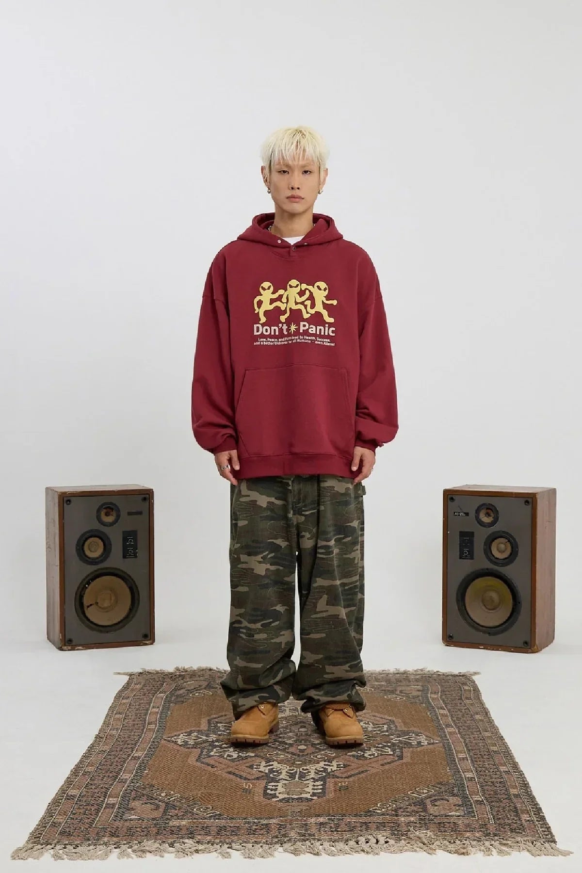 Young person wearing burgundy alien hoodie, camouflage pants, and boots standing on patterned rug between vintage speakers