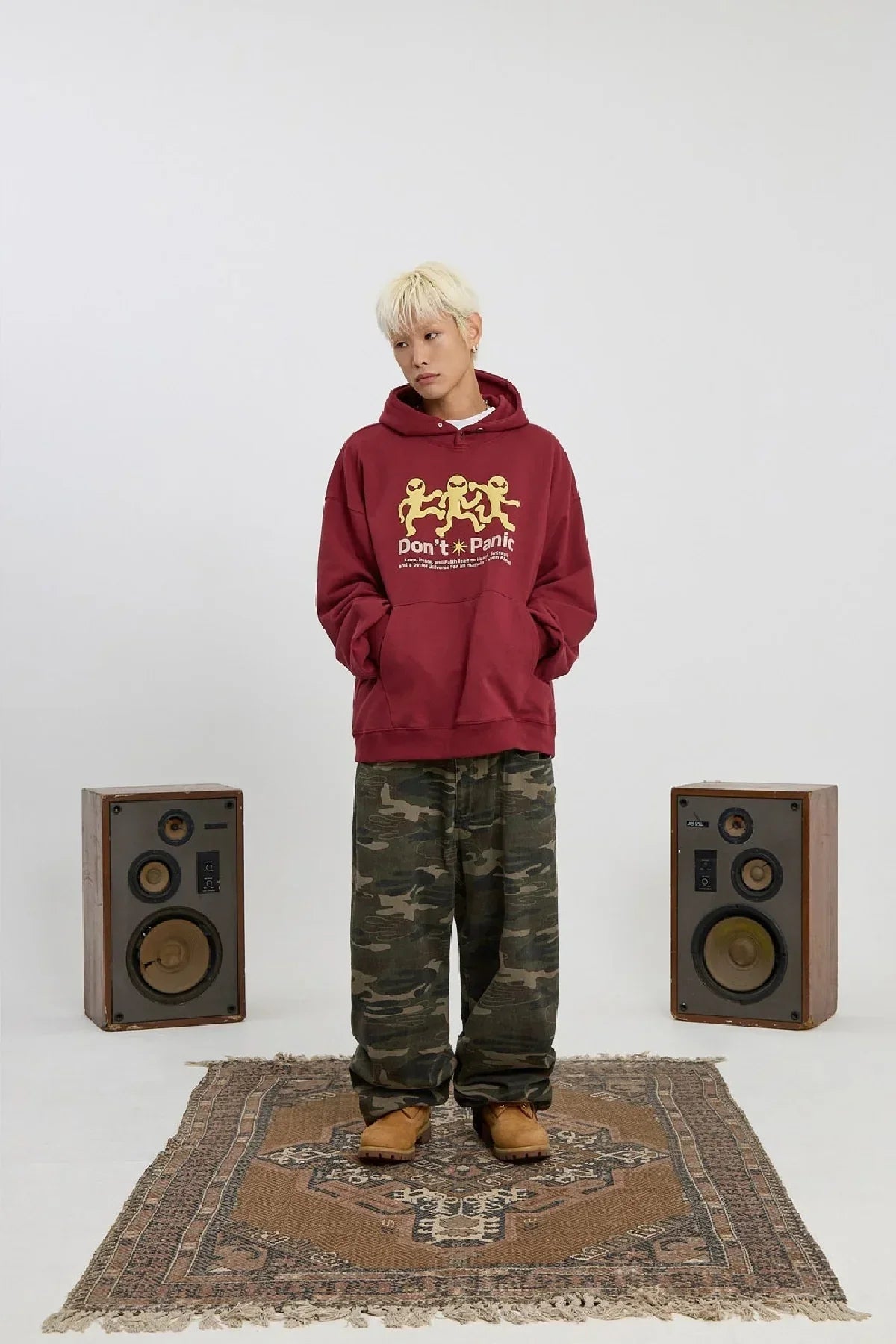 person wearing burgundy hoodie with three yellow aliens and text, camo pants, standing on patterned rug between two speakers