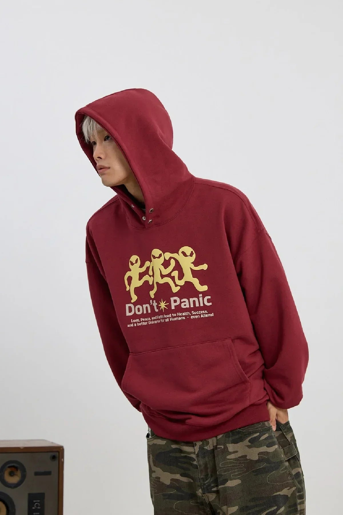 Young man wearing burgundy hoodie with yellow alien graphic and don't panic text, paired with camouflage pants