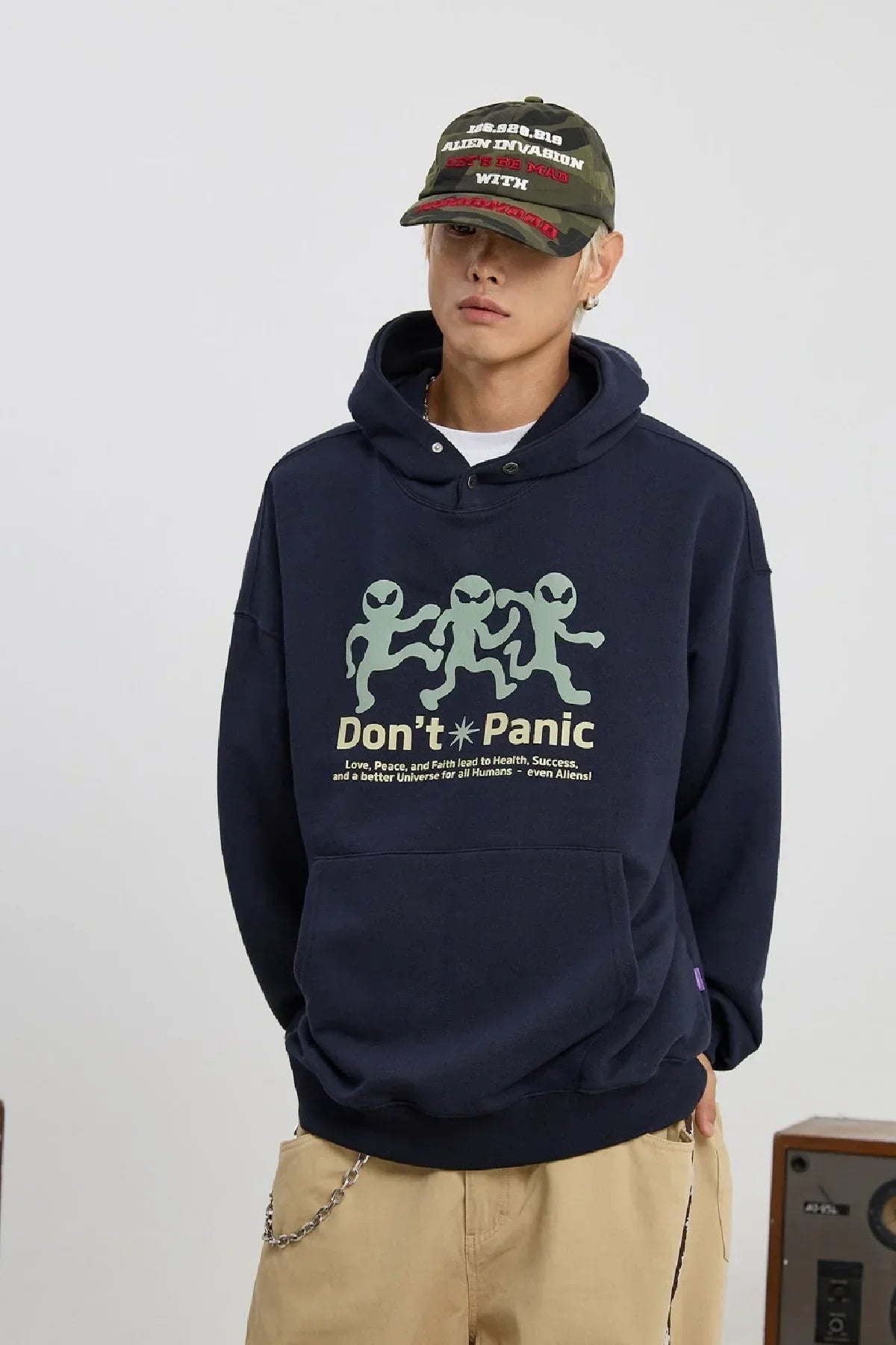 Man wearing navy hoodie with three green aliens and "Don't Panic" text, paired with camo cap and beige pants
