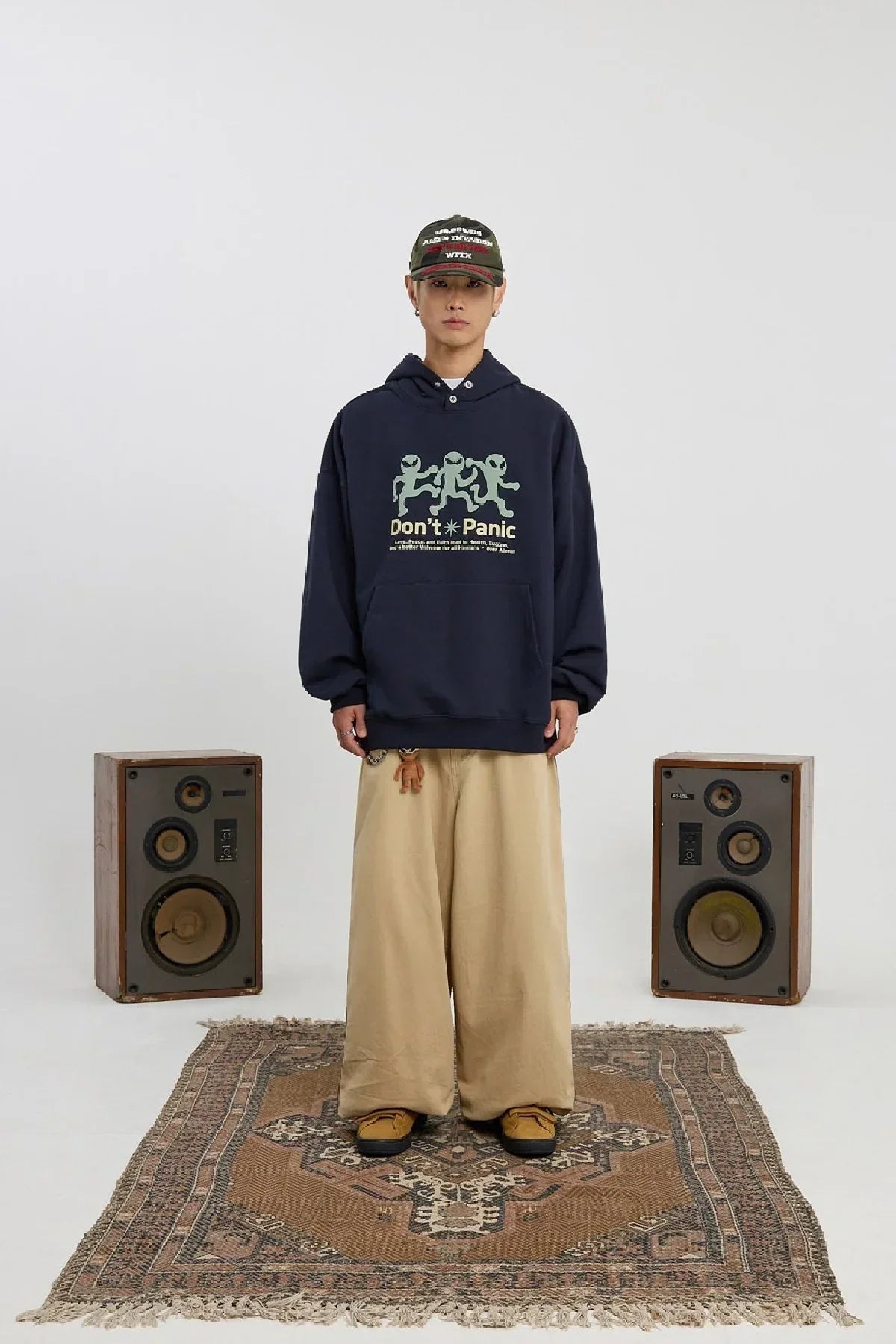 Model wearing navy alien graphic hoodie and beige wide-leg pants standing on patterned rug between two vintage speakers
