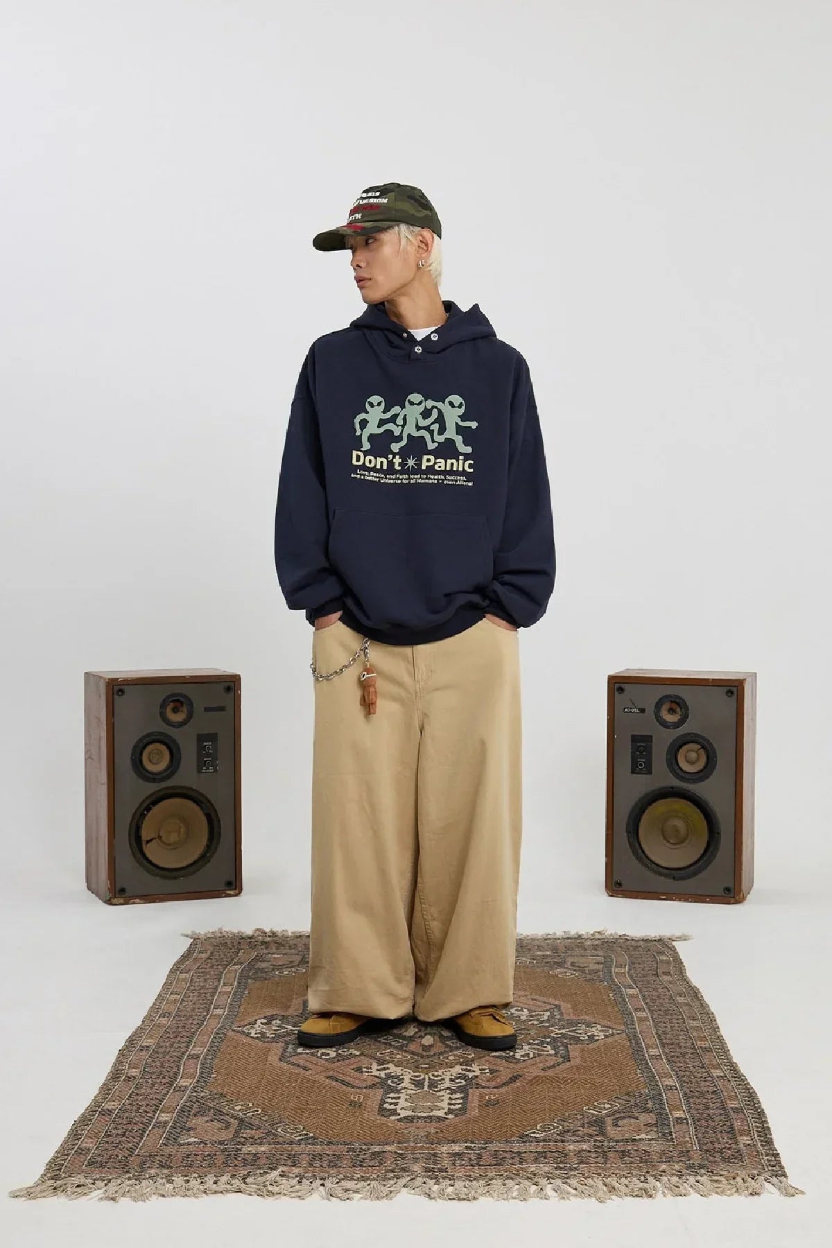 Person wearing navy alien-themed hoodie and wide beige pants standing on a patterned rug between vintage speakers