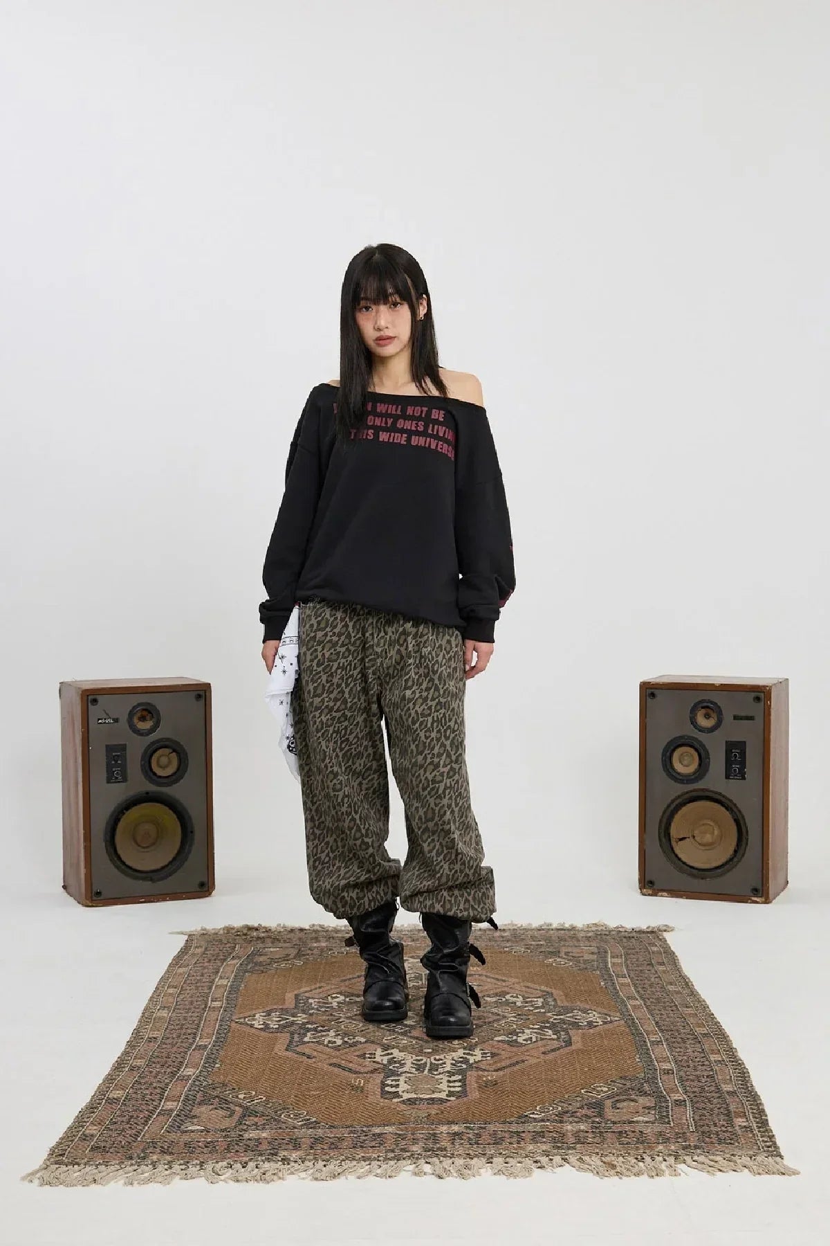 Model wearing black off-shoulder sweatshirt and leopard print pants standing on patterned rug between vintage speakers