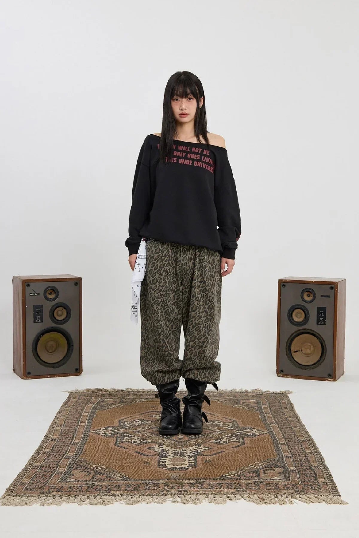Young woman in off-shoulder black sweatshirt and leopard print pants standing on patterned rug between vintage speakers
