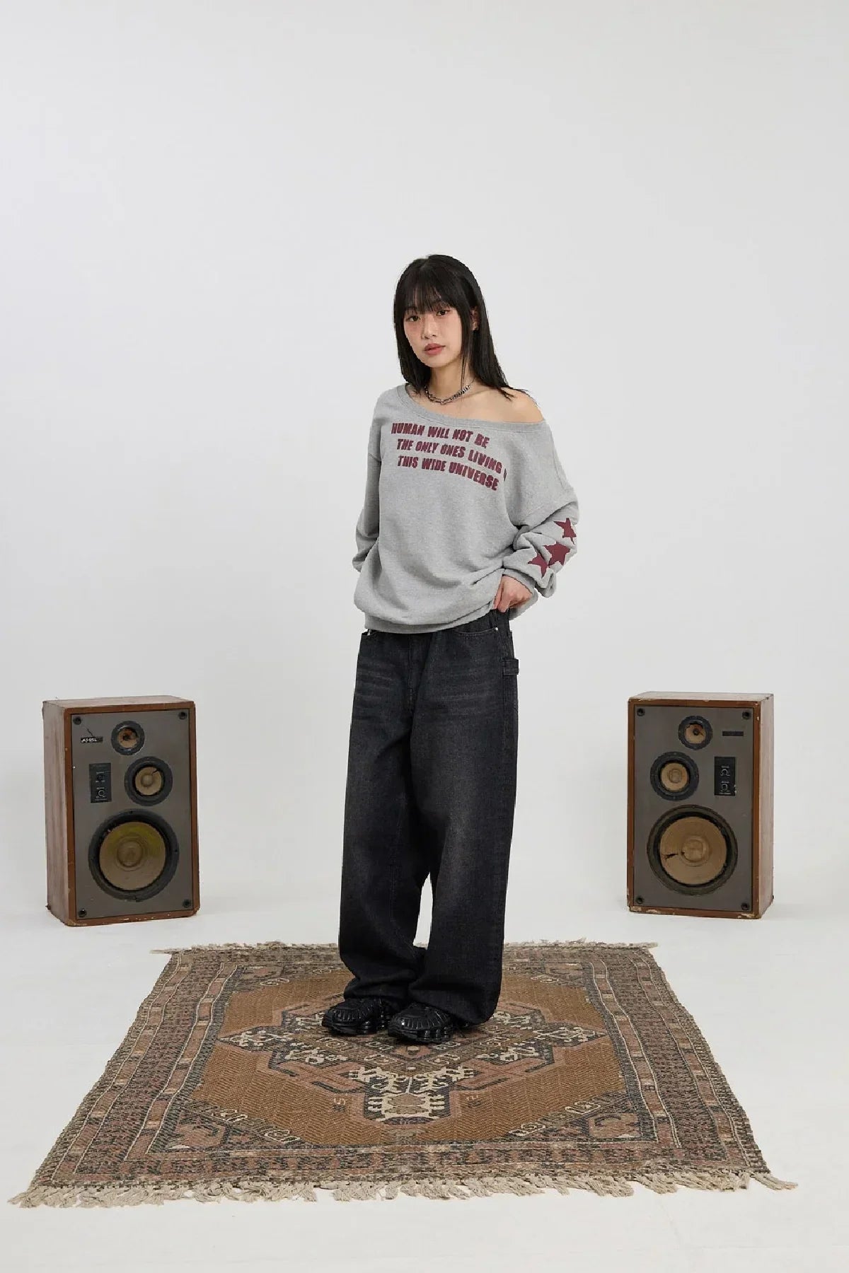 Woman wearing grey off-shoulder sweatshirt with red stars and text, black wide jeans, standing on patterned rug between vintage speakers