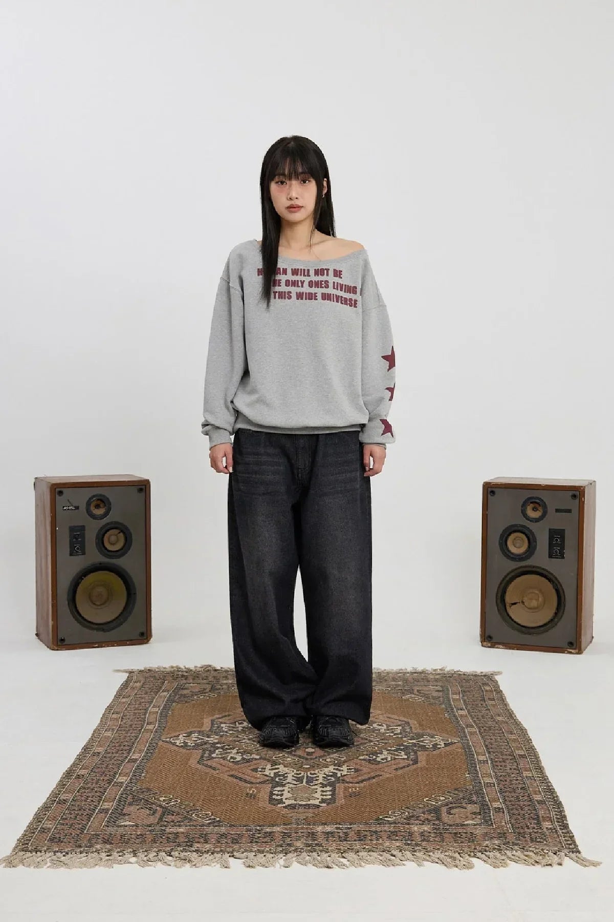 Woman standing on patterned rug wearing grey off-shoulder sweatshirt with red text and stars, black wide pants