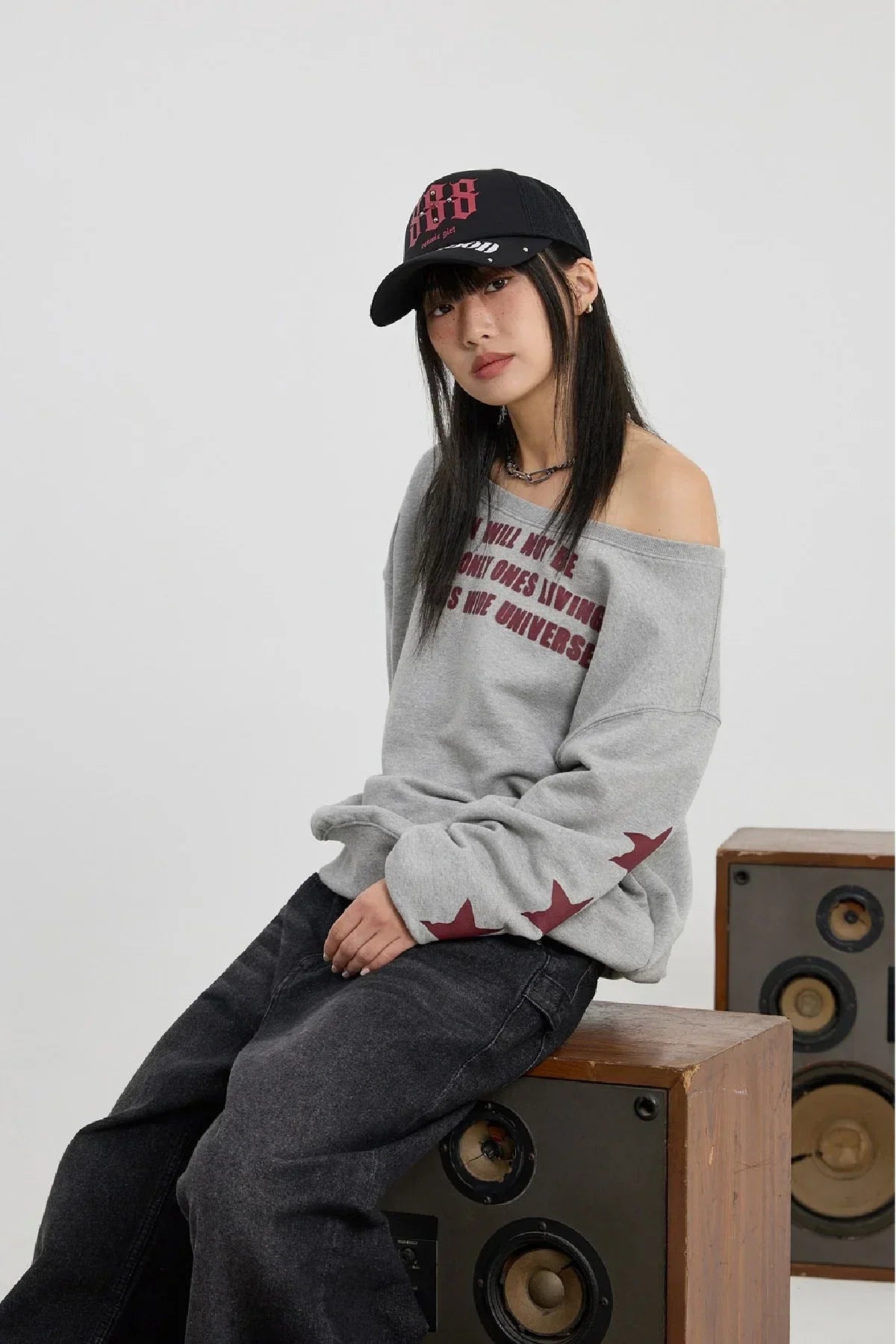 Woman wearing grey off-shoulder sweatshirt with star details and black cap, sitting on vintage speaker