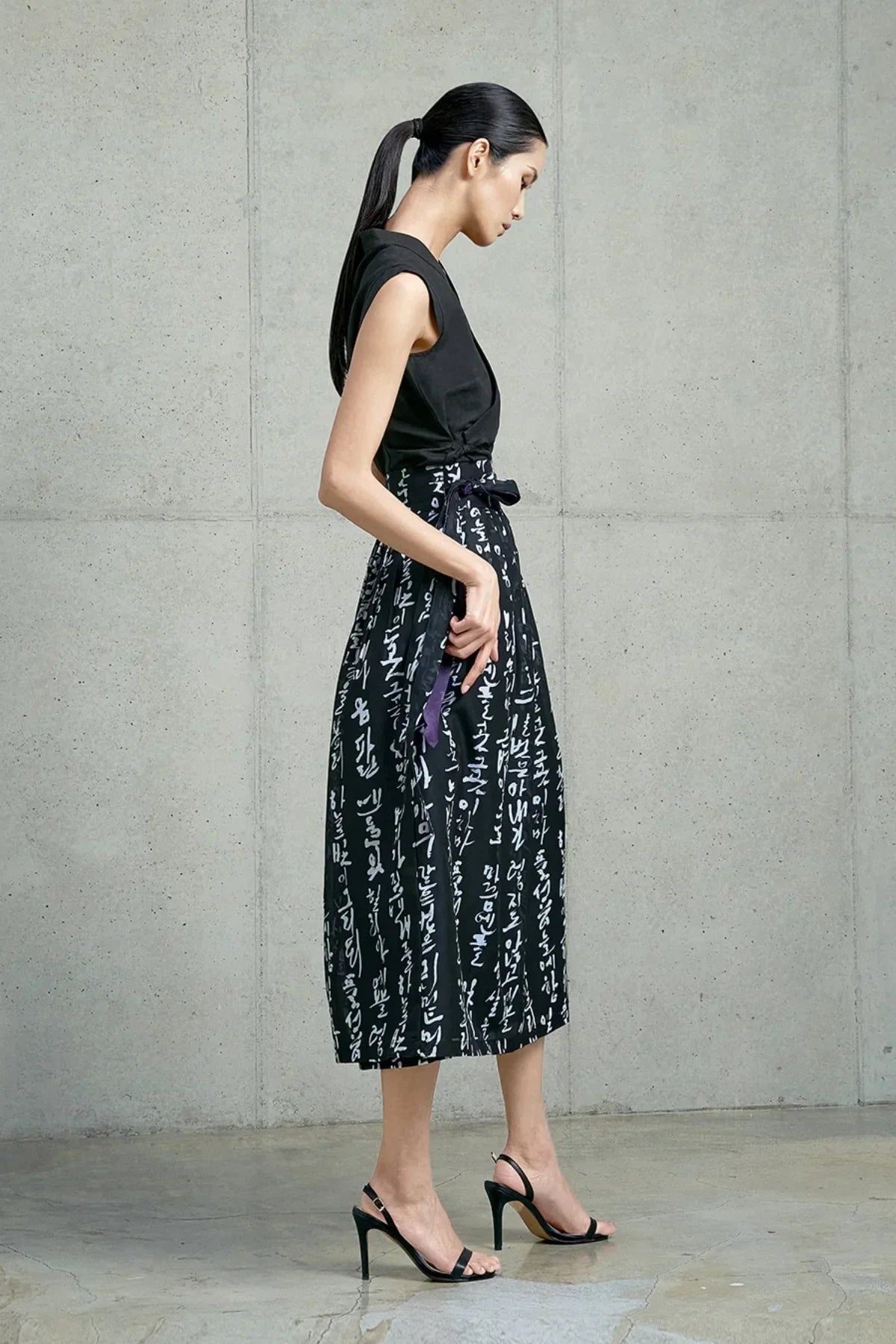 Woman in black sleeveless top and black midi skirt with white Hangeul script and purple ribbon, standing against concrete wall