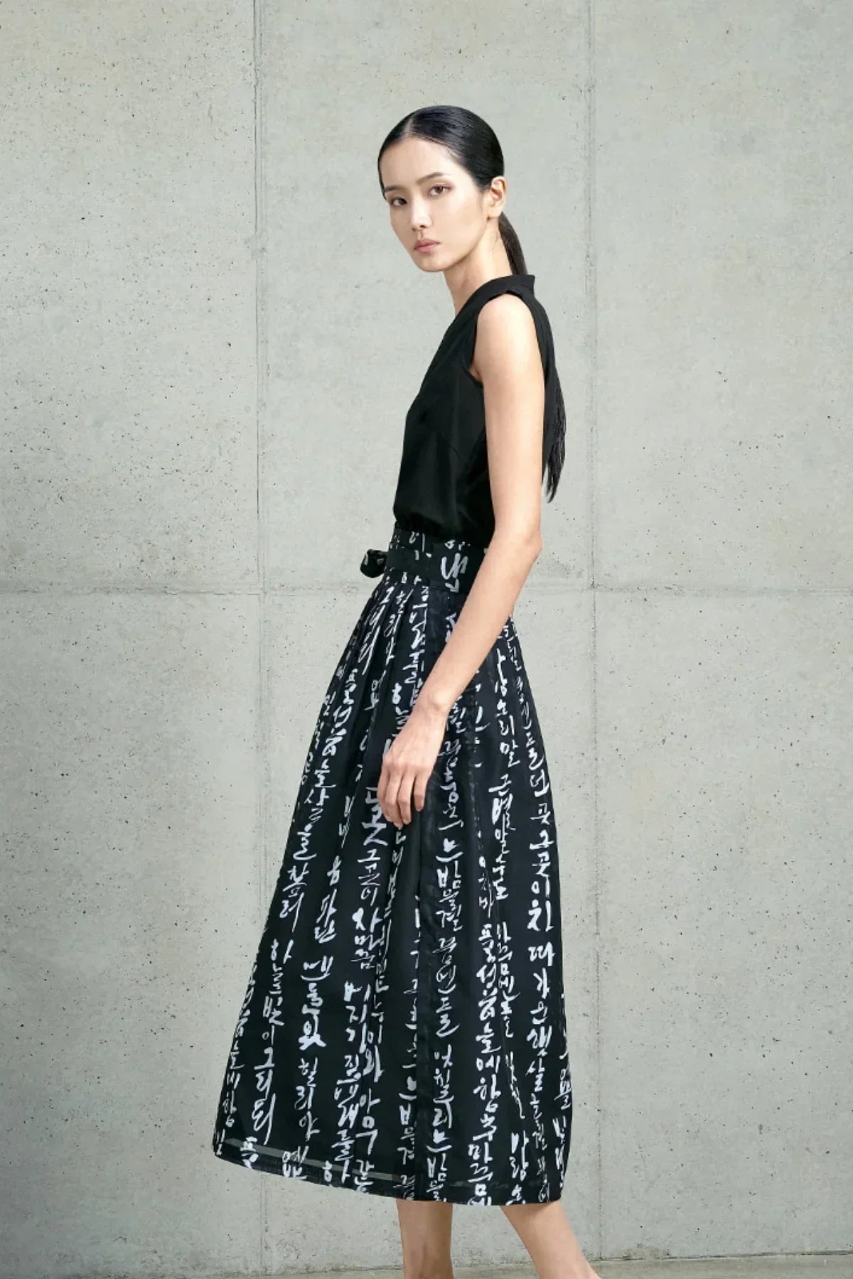 Woman in black sleeveless top and black skirt with white traditional Hangeul print standing against concrete wall