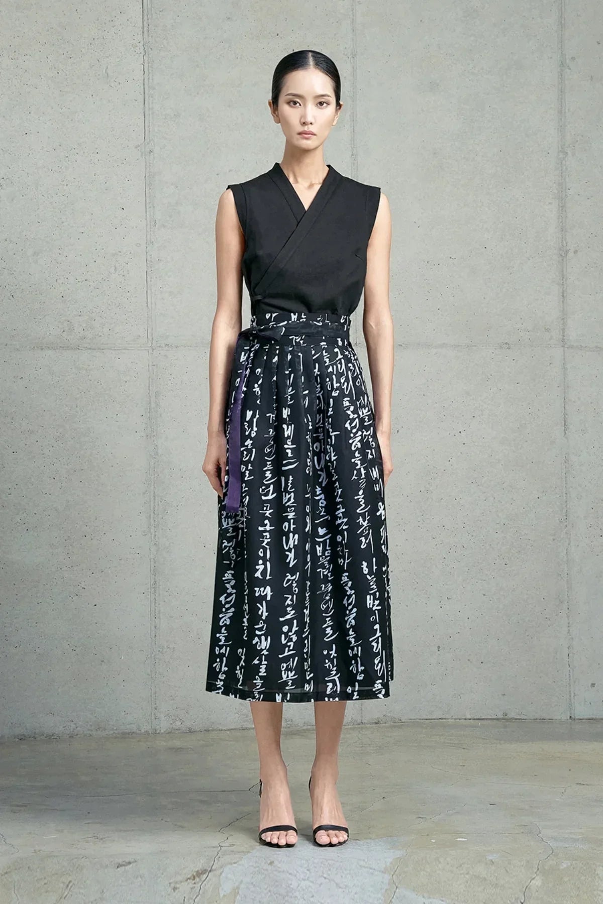 Asian woman wearing black sleeveless top and midi skirt with white Hangeul script, standing in minimalist concrete room