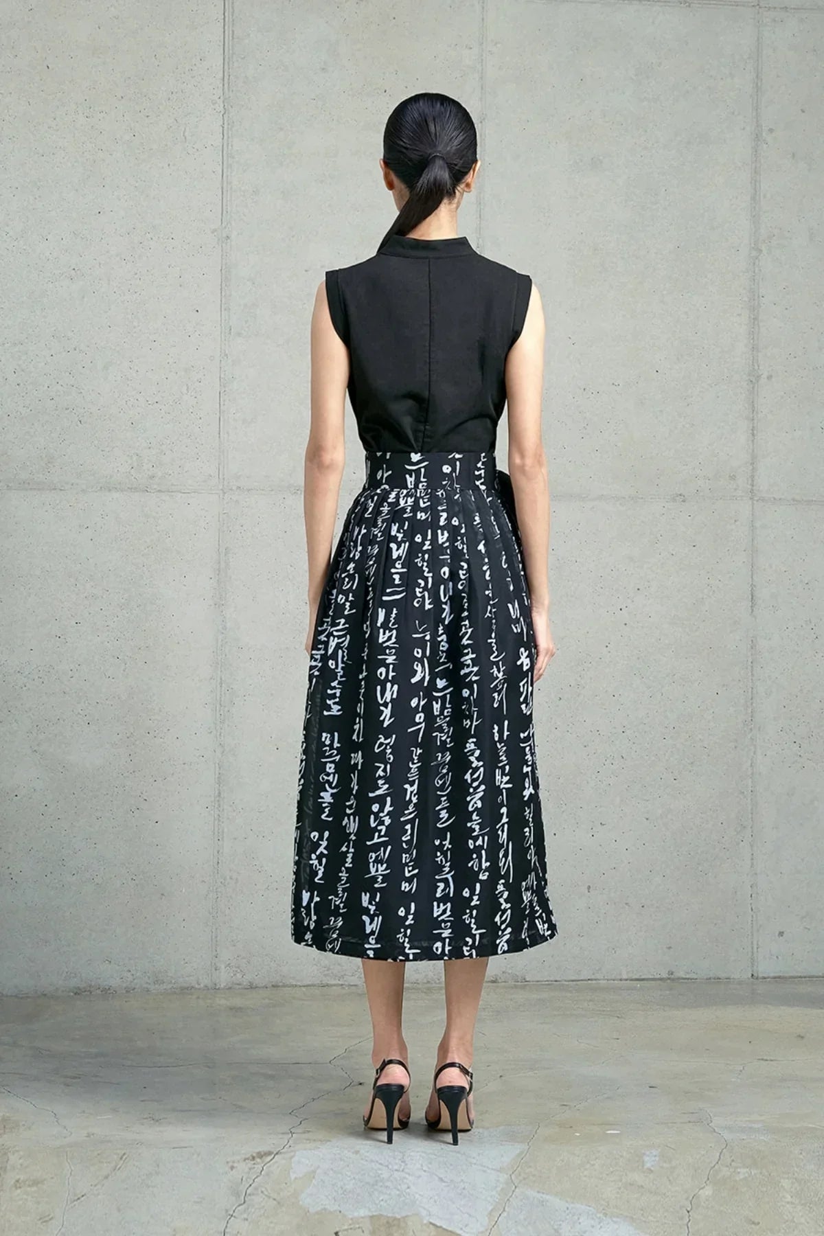 Woman wearing a black sleeveless top and a midi skirt with white traditional Hangul script, standing against a concrete wall