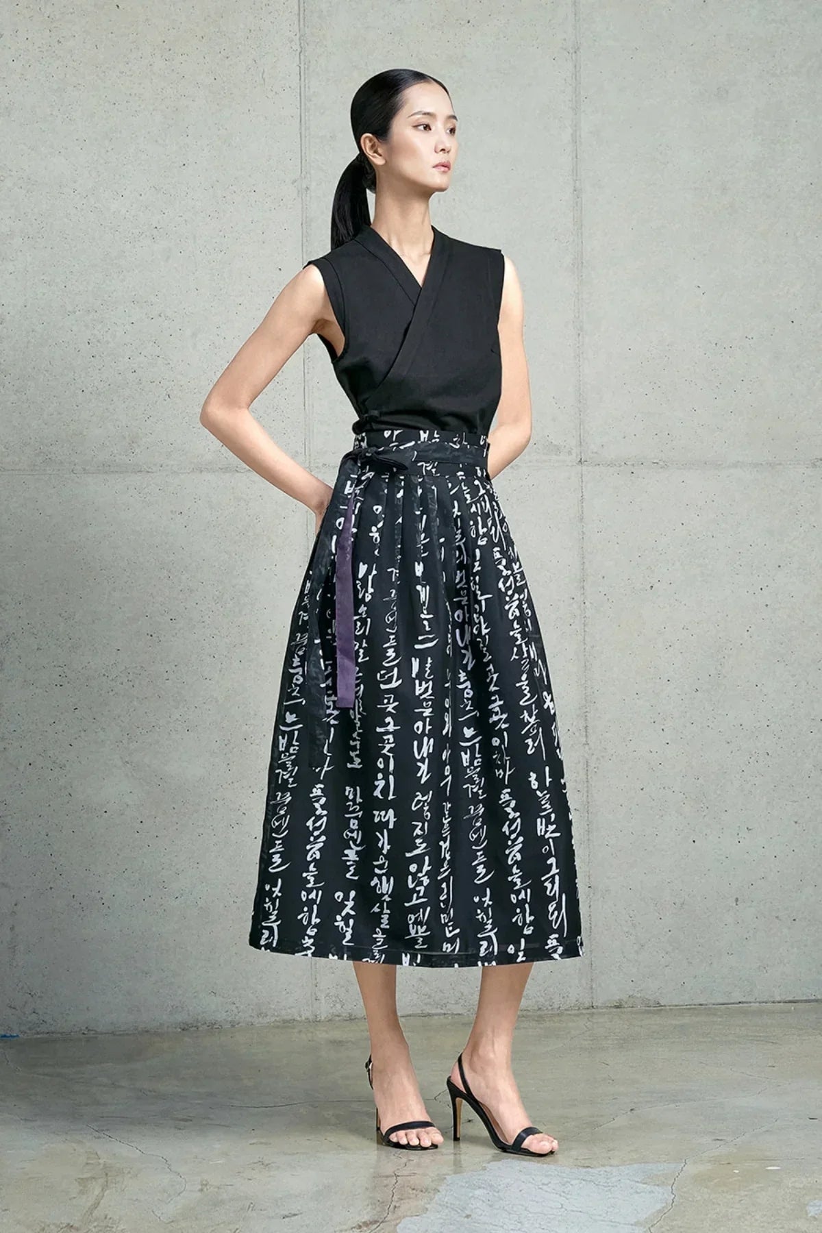 Woman in black sleeveless top and black pleated skirt with white Hangeul script, standing against concrete wall
