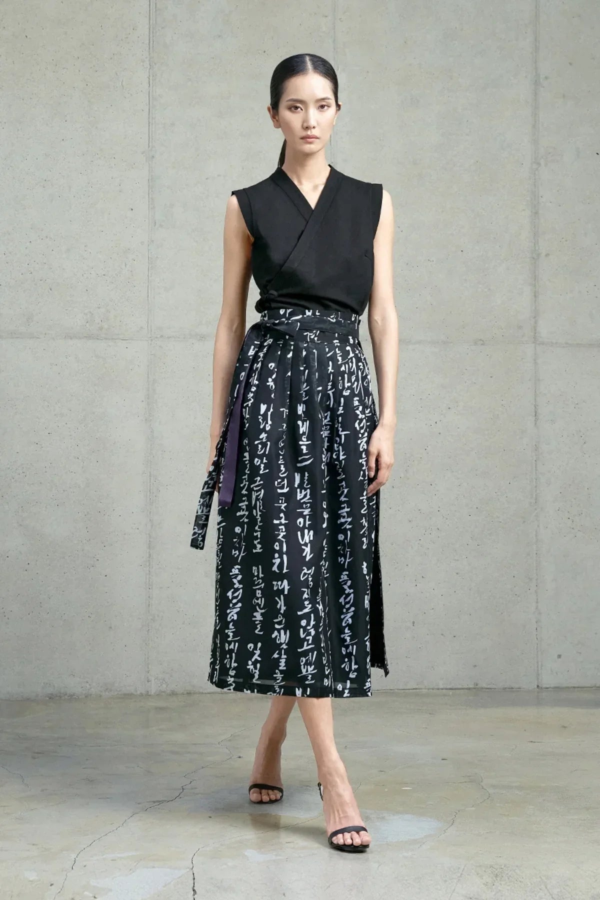 Woman wearing a black sleeveless top and a black wrap skirt with white Hangeul script, standing against a concrete wall