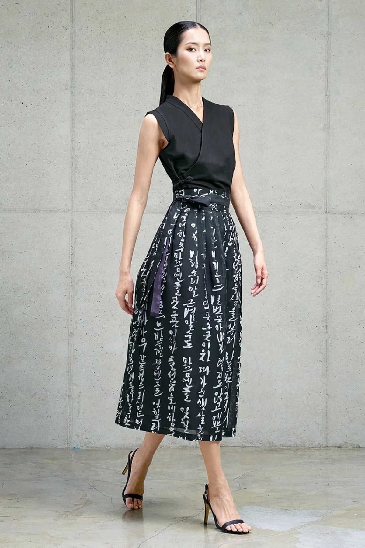 Model wearing a black sleeveless top with a traditional Hangeul printed black waist skirt and black heels on concrete floor