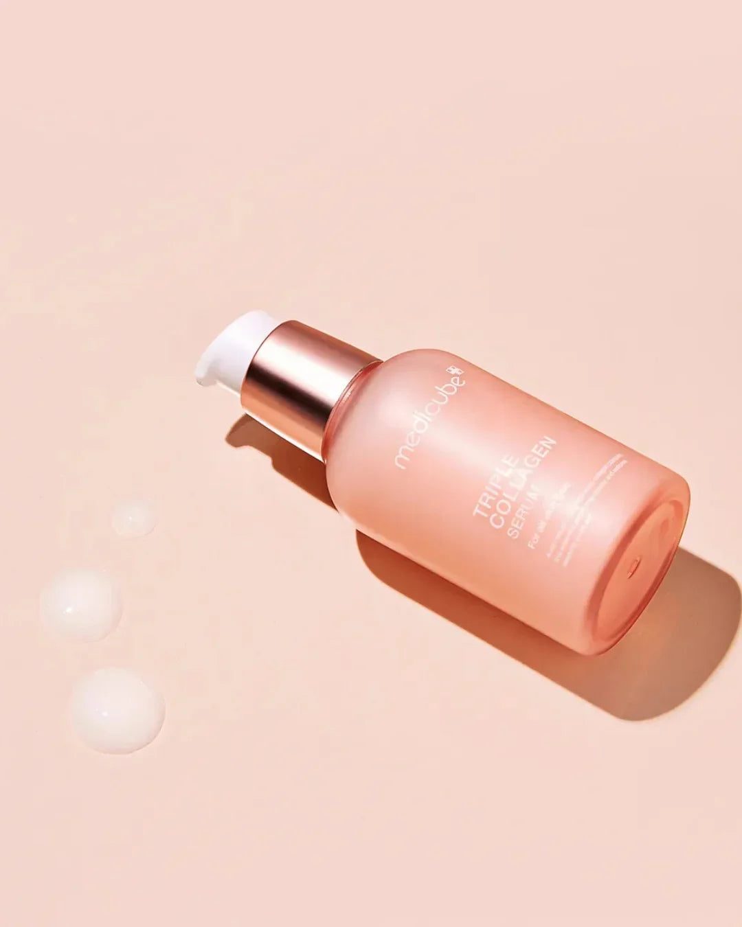 Medicube Triple Collagen Serum 4.0 in pink frosted bottle with pump on peach background with serum droplets