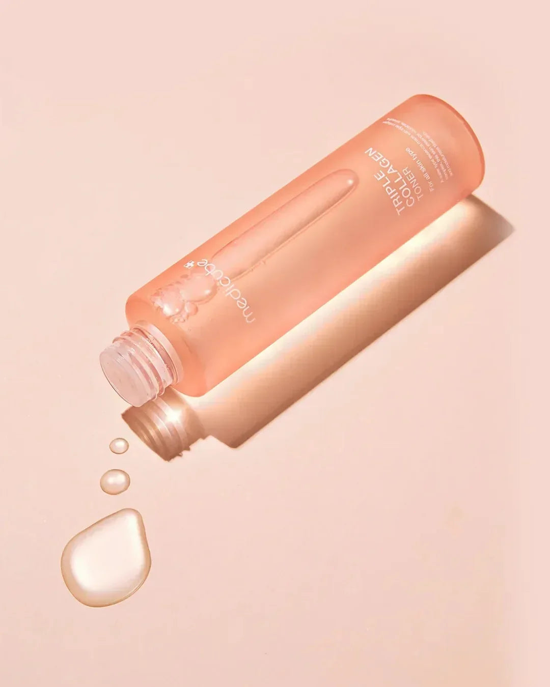 Open pink bottle of Medicube Triple Collagen Toner 4.0 with liquid drops on light pink background
