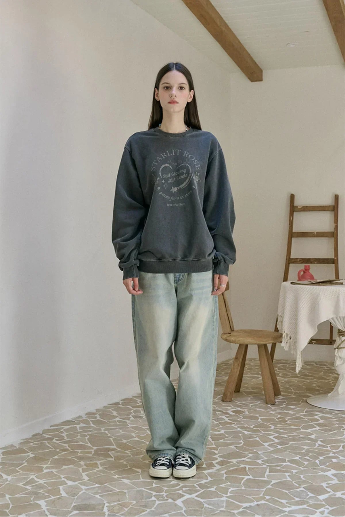 Woman wearing a smoke black Star Lit Rose graphic sweatshirt and light washed jeans in a minimalist room