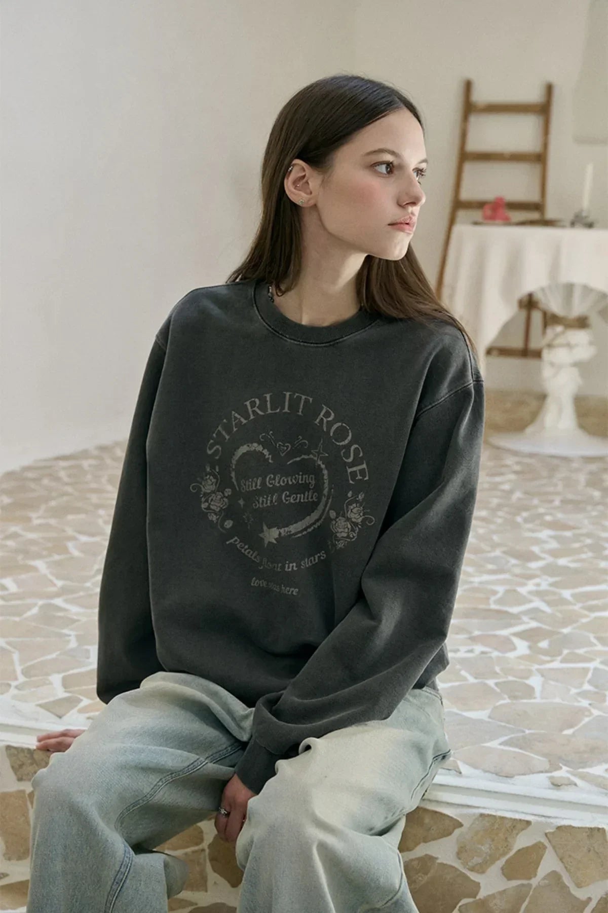 Young woman wearing a smoke black Starlit Rose sweatshirt with graphic design, sitting indoors on tiled floor