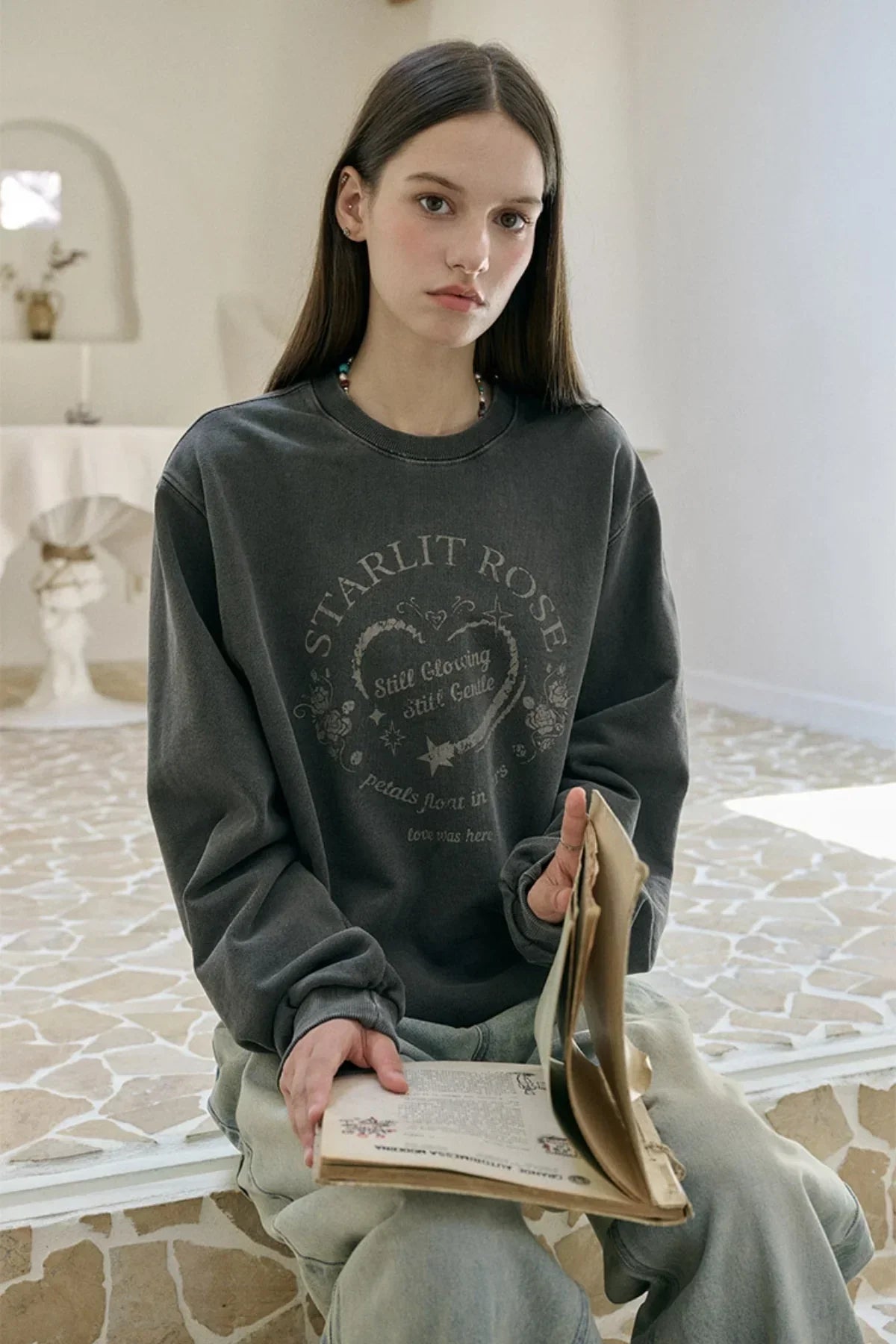 Young woman wearing a smoke black Starlit Rose sweatshirt sitting indoors reading a vintage book