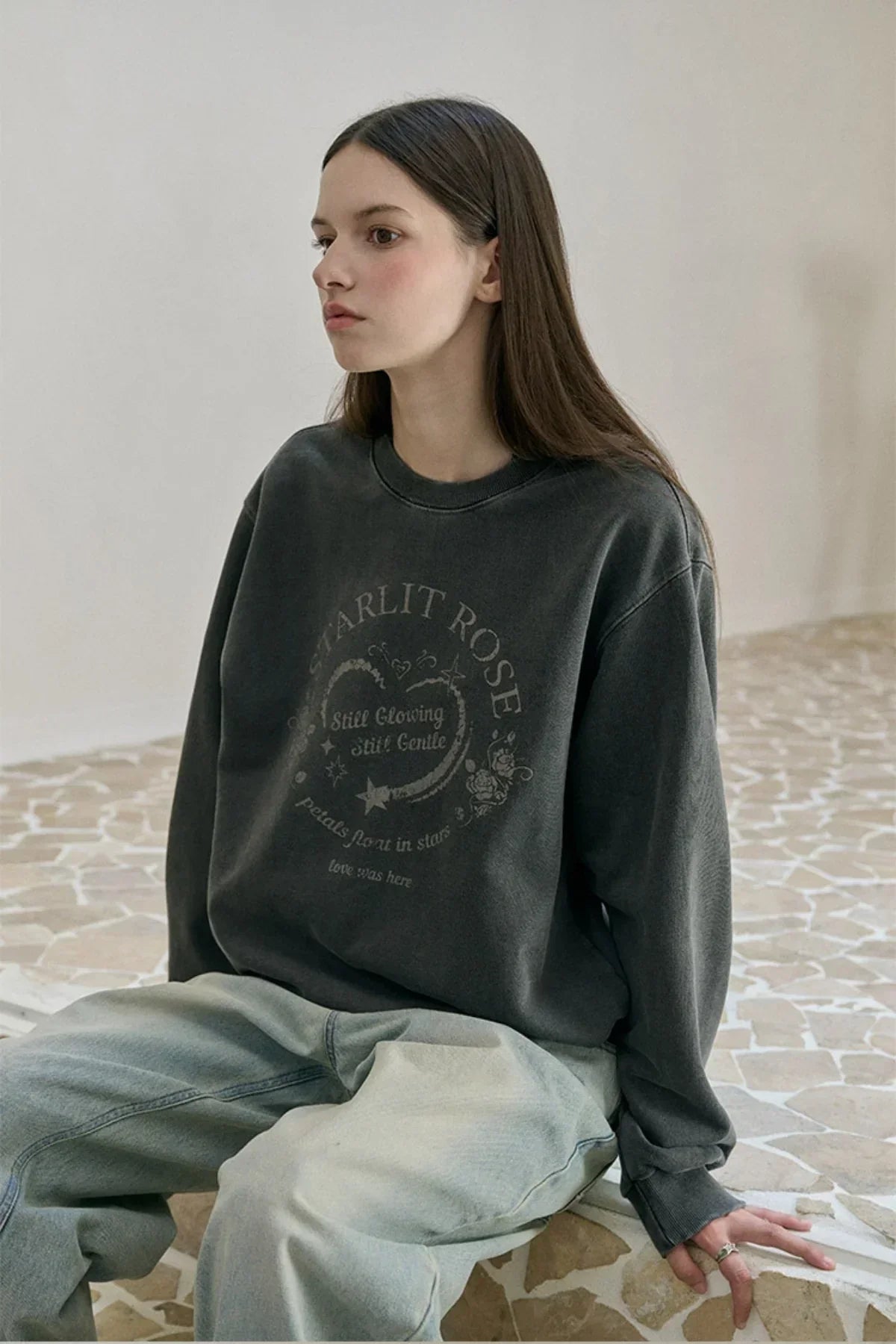 Young woman in dark grey graphic sweatshirt and light washed jeans sitting on stone floor indoors