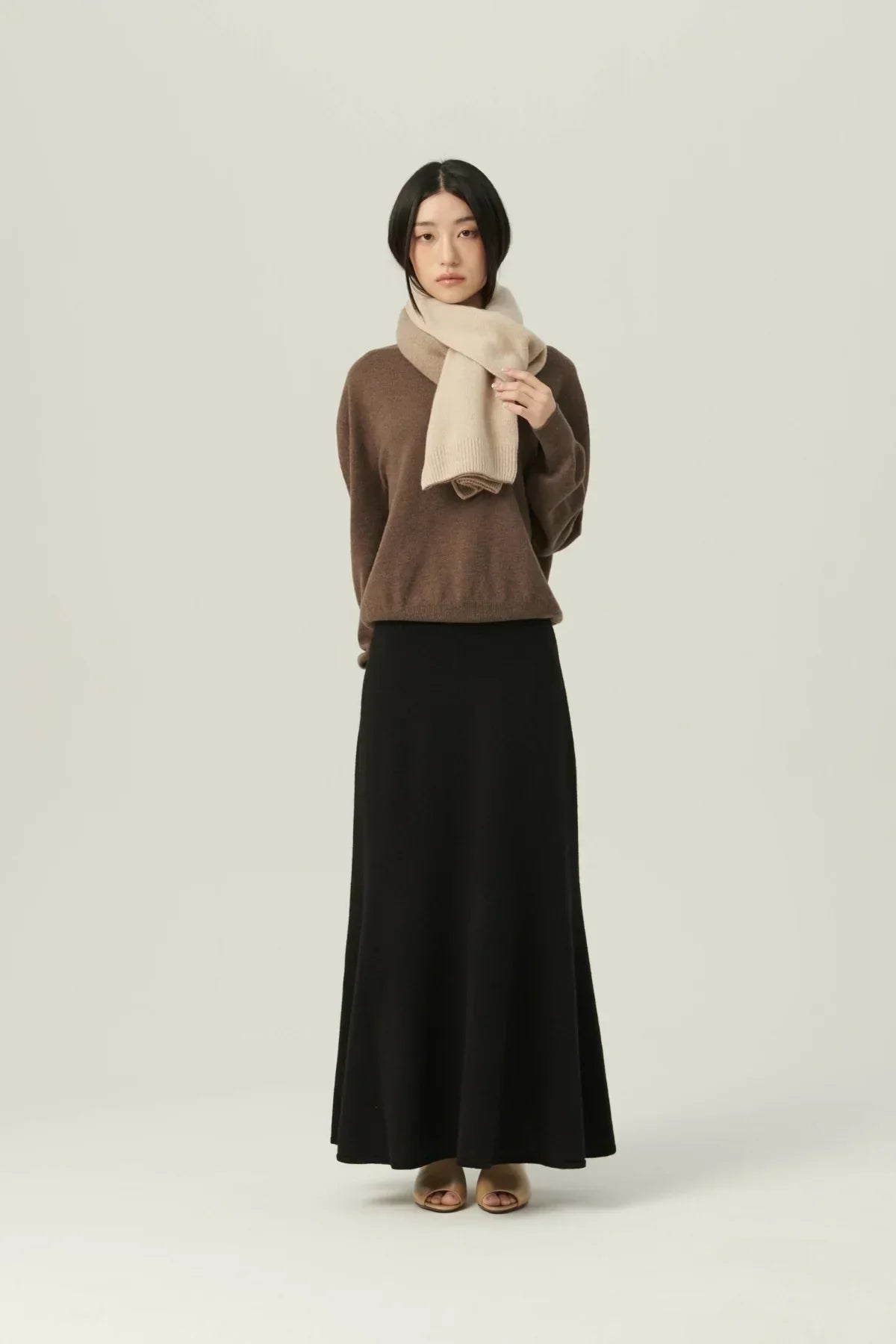 Woman wearing beige two-tone cashmere muffler, brown sweater, and long black skirt on plain background