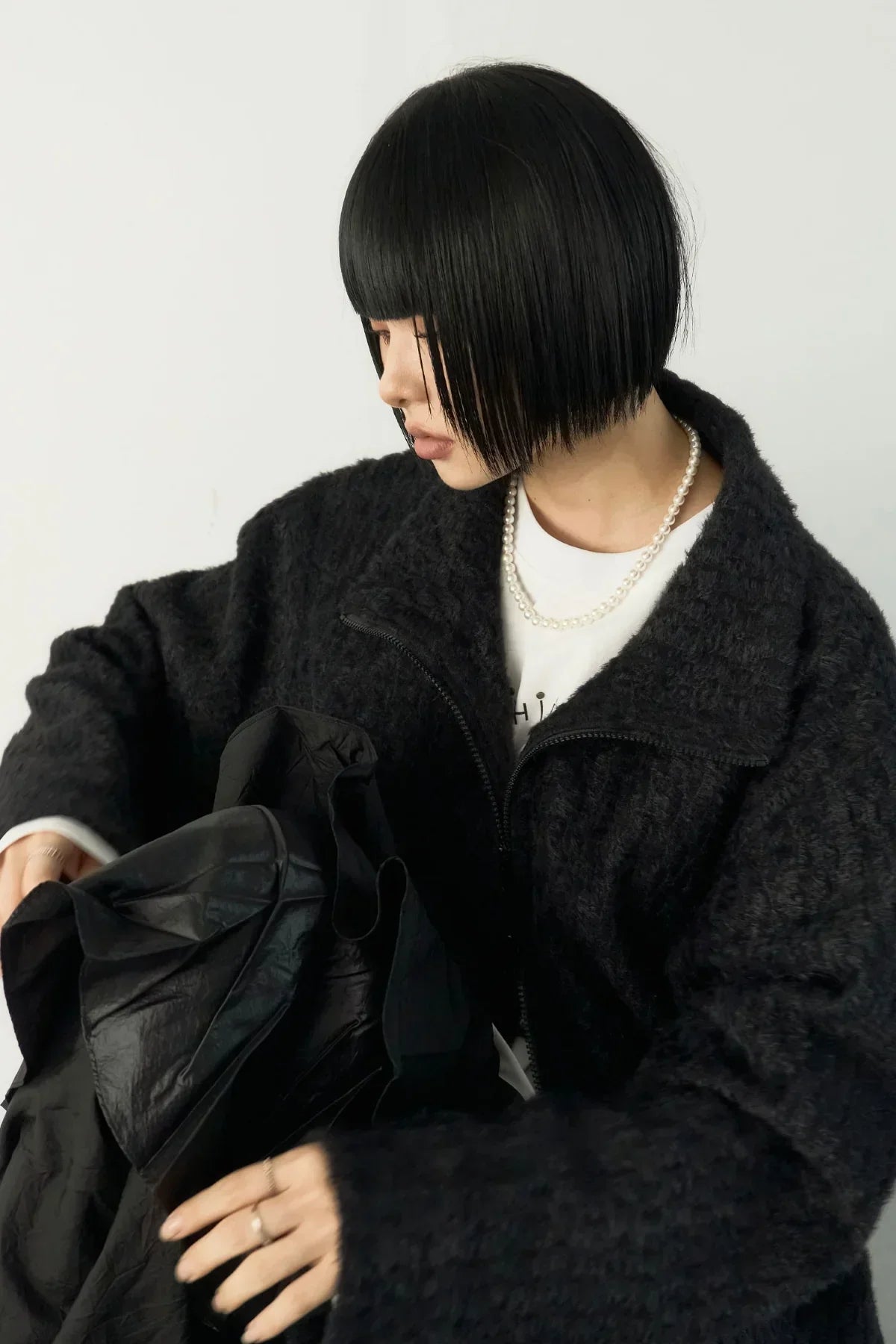 Woman with short black hair wearing a textured black jacket and pearl necklace holding a black garment