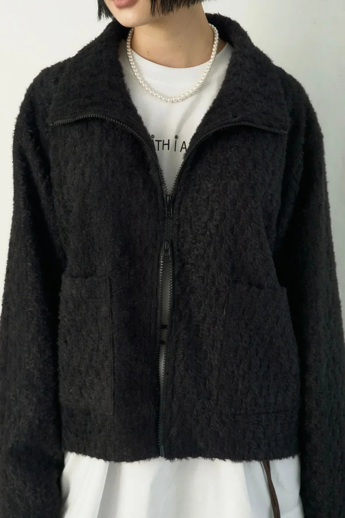 Close-up of woman wearing textured black zip-up jacket, white shirt, and pearl necklace